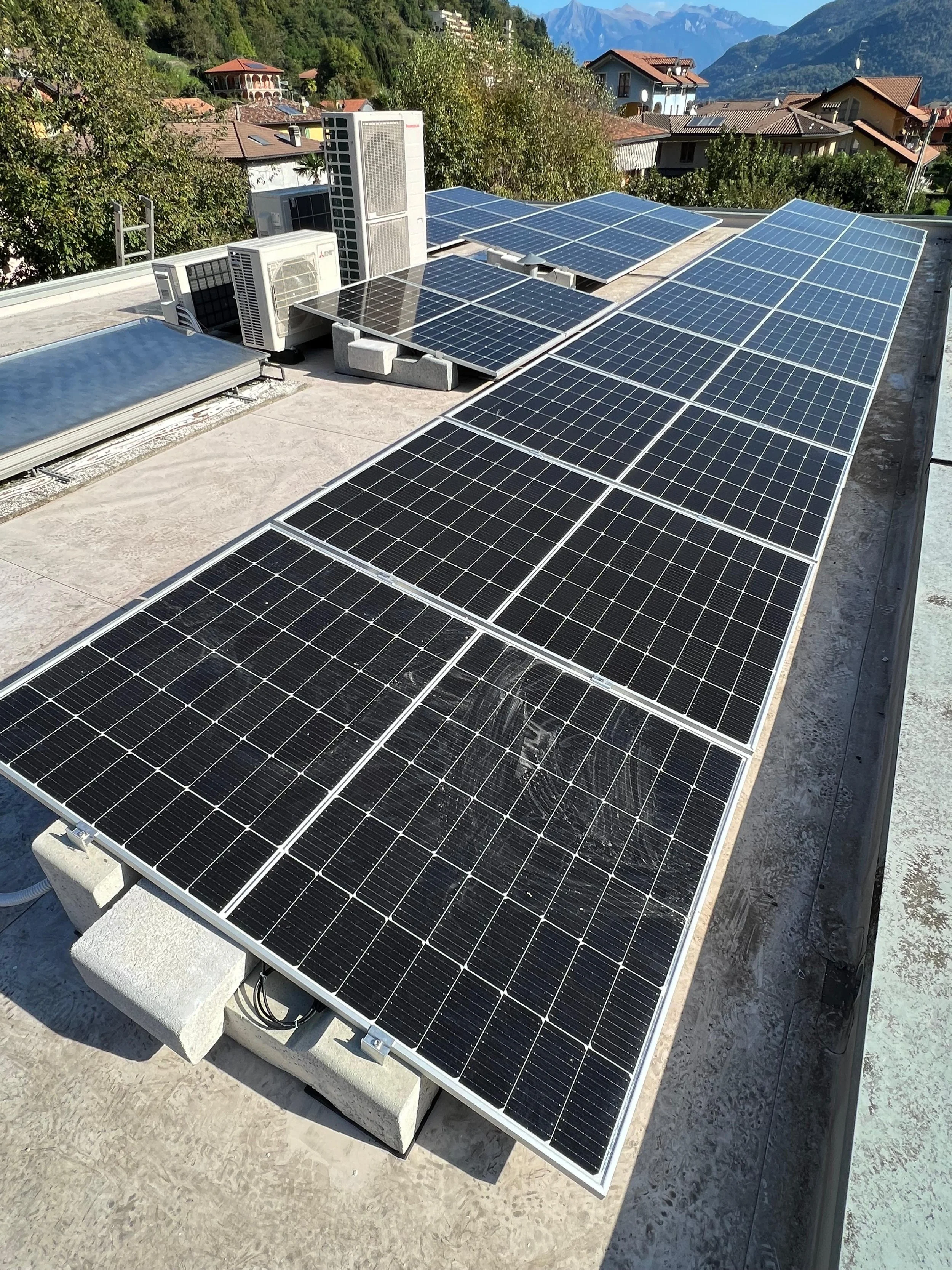 Pannelli solari installati sul tetto di un edificio, con arredi e condizionatori visibili, in un'area montuosa con case e alberi in background.