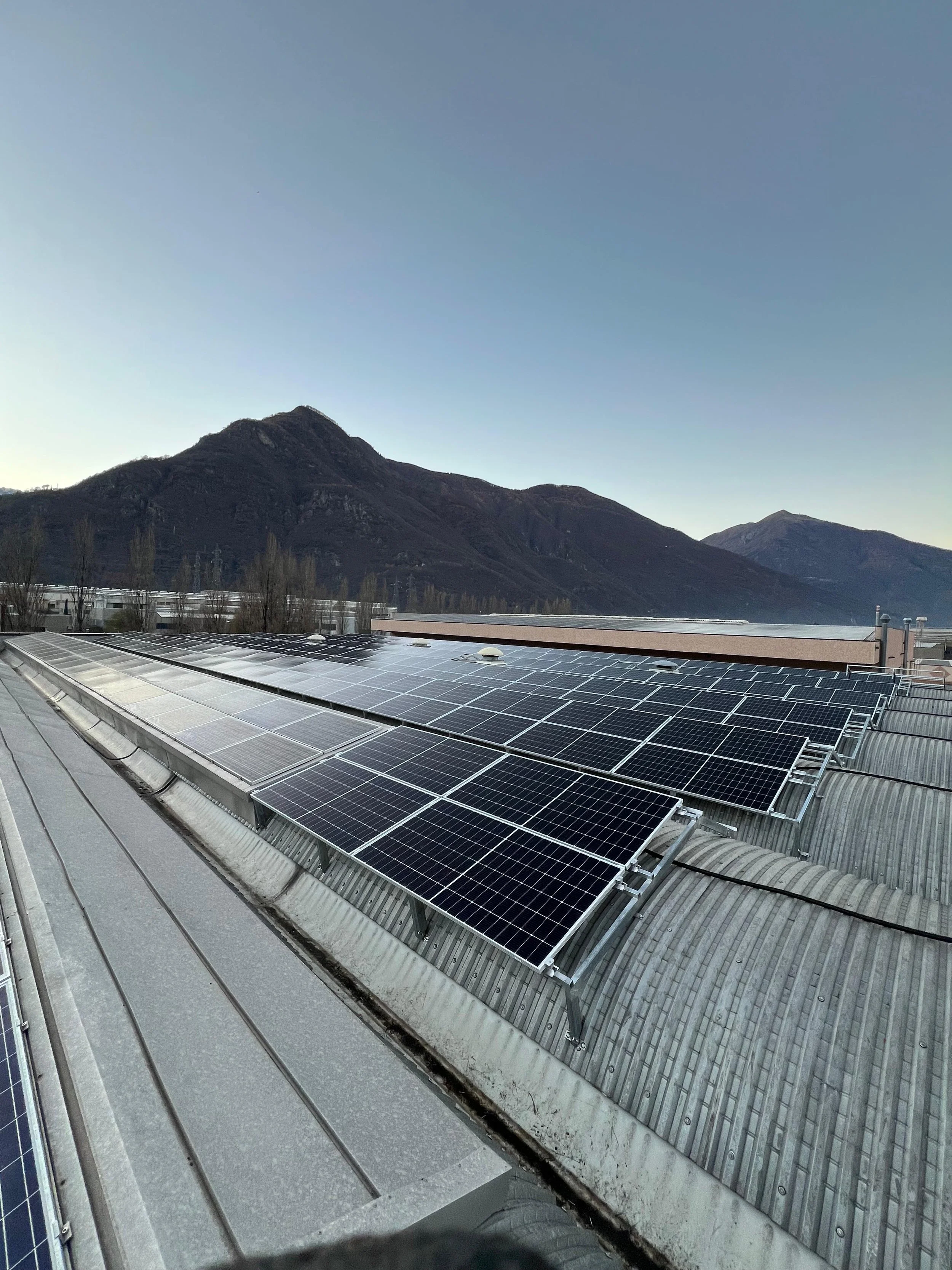 Panoramica di pannelli solari installati su un tetto industriale con montagne sullo sfondo e un cielo chiaro.