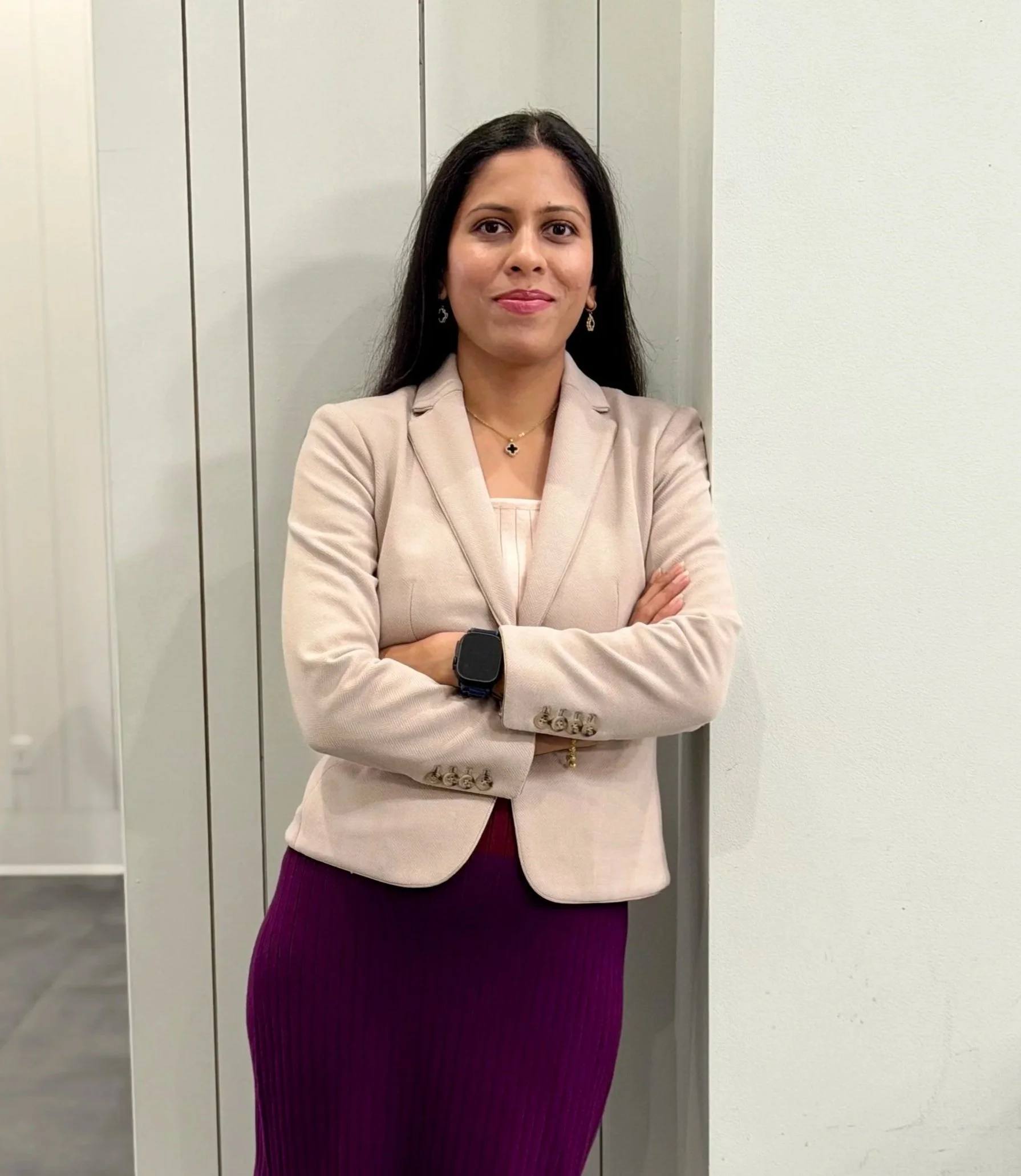 A woman with long dark hair standing with arms crossed in front of an elevator door against a white wall, wearing a beige blazer, purple skirt, earrings, necklace, and smartwatch.