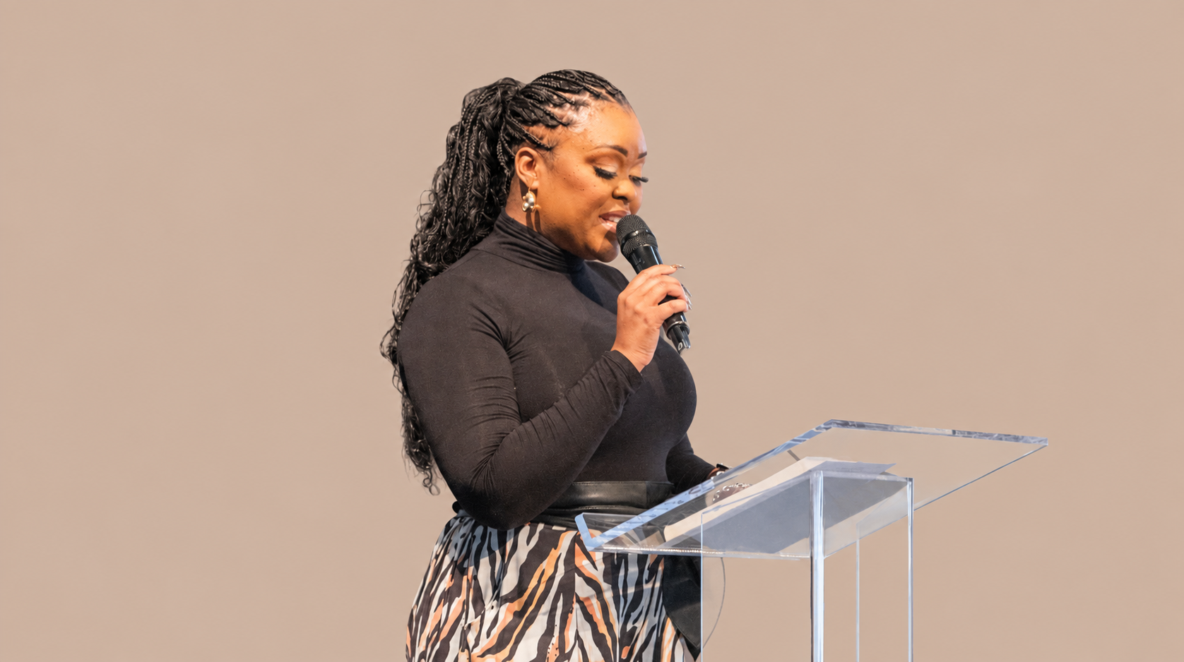 A woman with long braided hair, wearing a black long-sleeve top and patterned skirt, standing at a clear podium and speaking into a microphone.