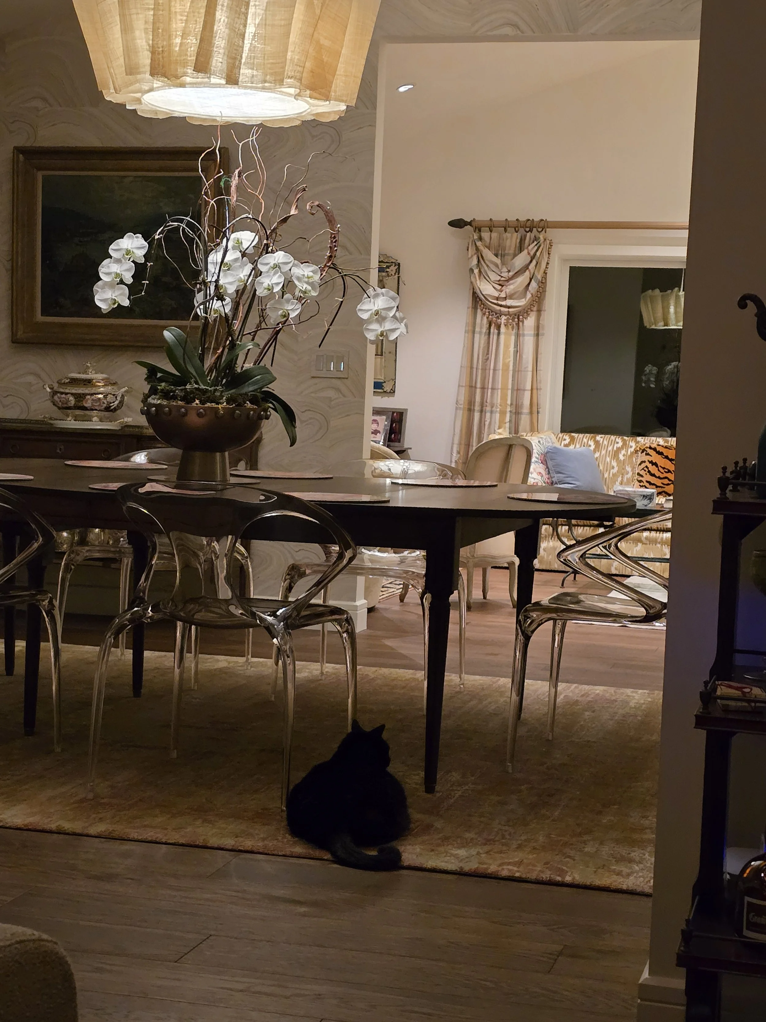 A dining room with a table and chairs, a floral centerpiece, wall art, and window with curtains. A black cat is sitting on a rug under the table.