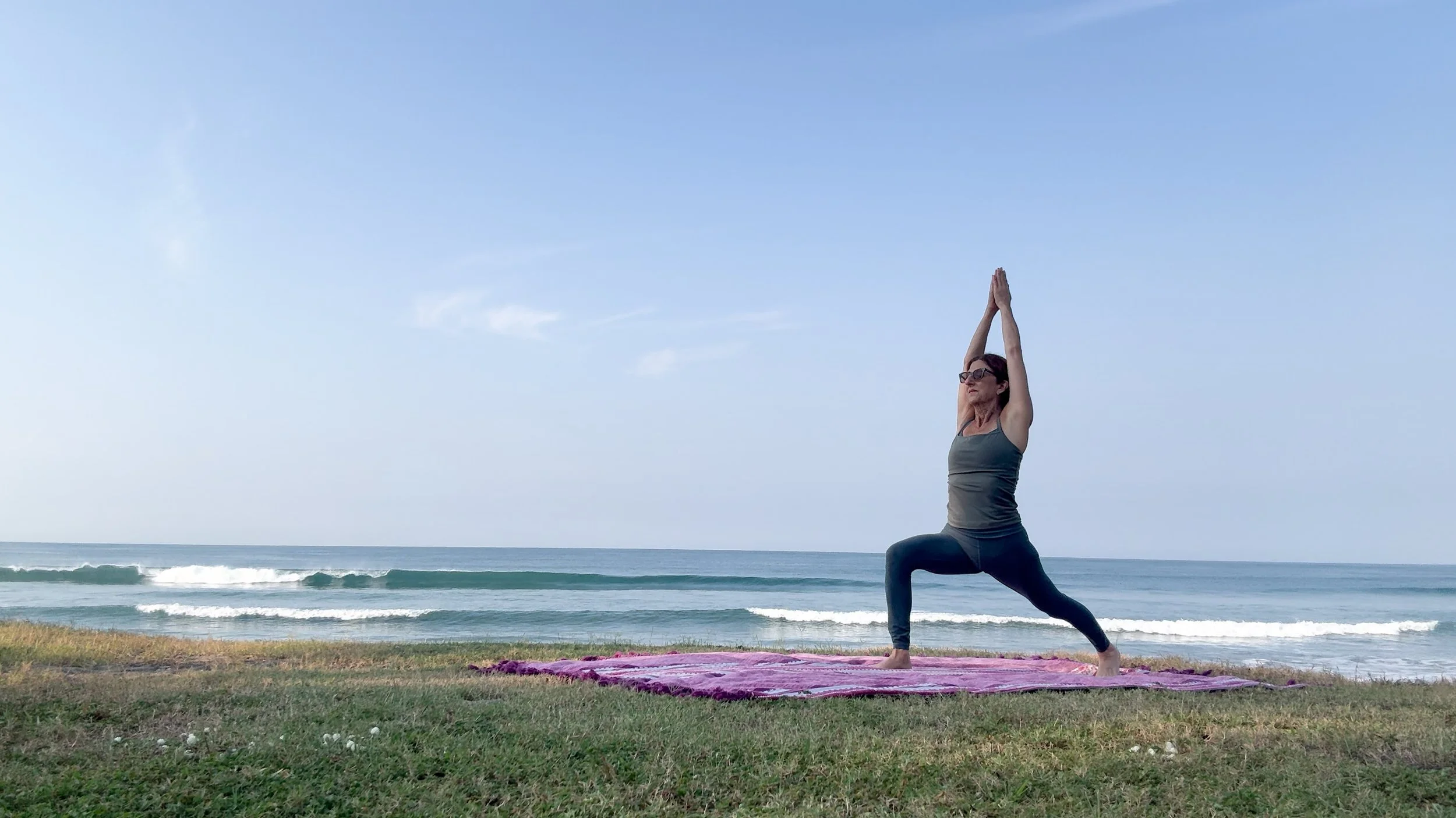 Carla practicing yoga by the ocean with her arms raised overhead.