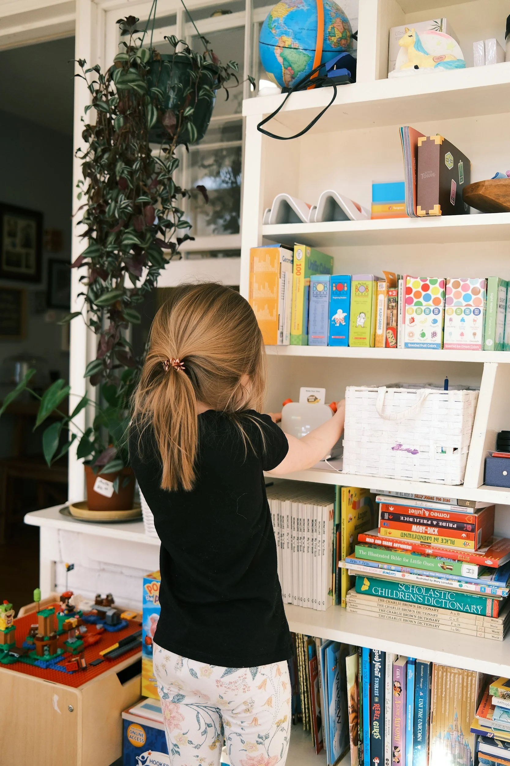 A young girl with light brown hair tied in a ponytail, wearing a black shirt and pastel-colored pajama pants with floral pattern, browsing books in a library or bookstore. She stands in front of white shelving filled with children's books and other educational items, with a large potted plant and toys nearby.