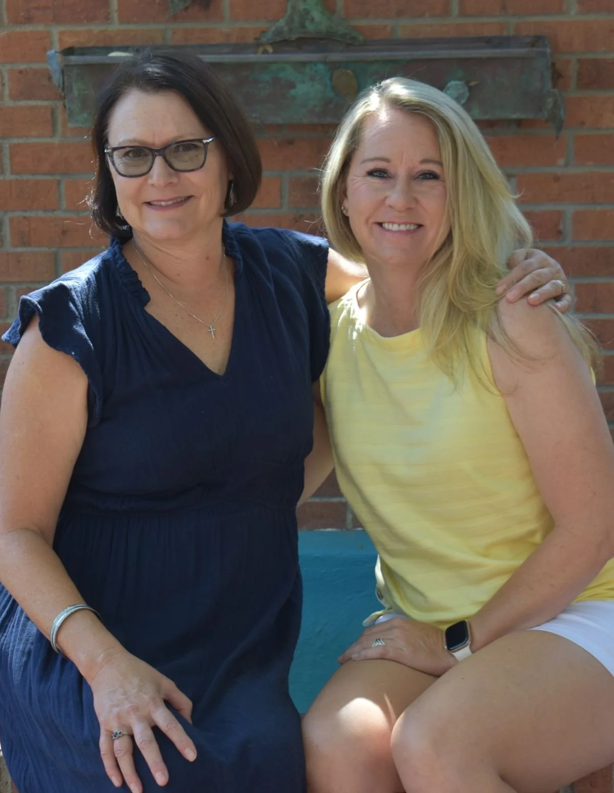 Two women sitting close together outdoors, one wearing glasses and a navy blue dress, the other in a yellow tank top, smiling at the camera with a brick wall behind them.