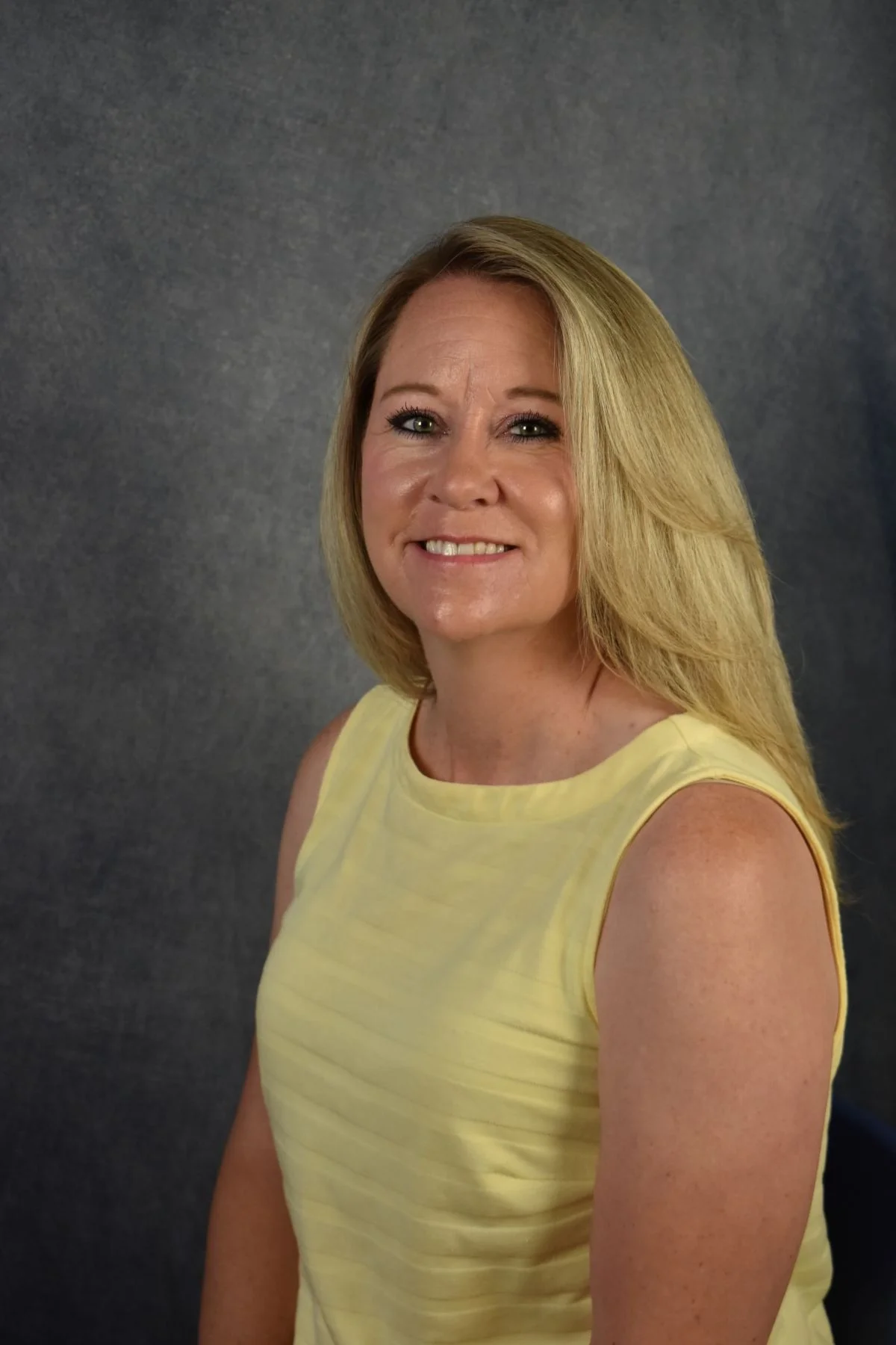 A smiling woman with blonde hair wearing a sleeveless yellow top standing against a dark grey background.