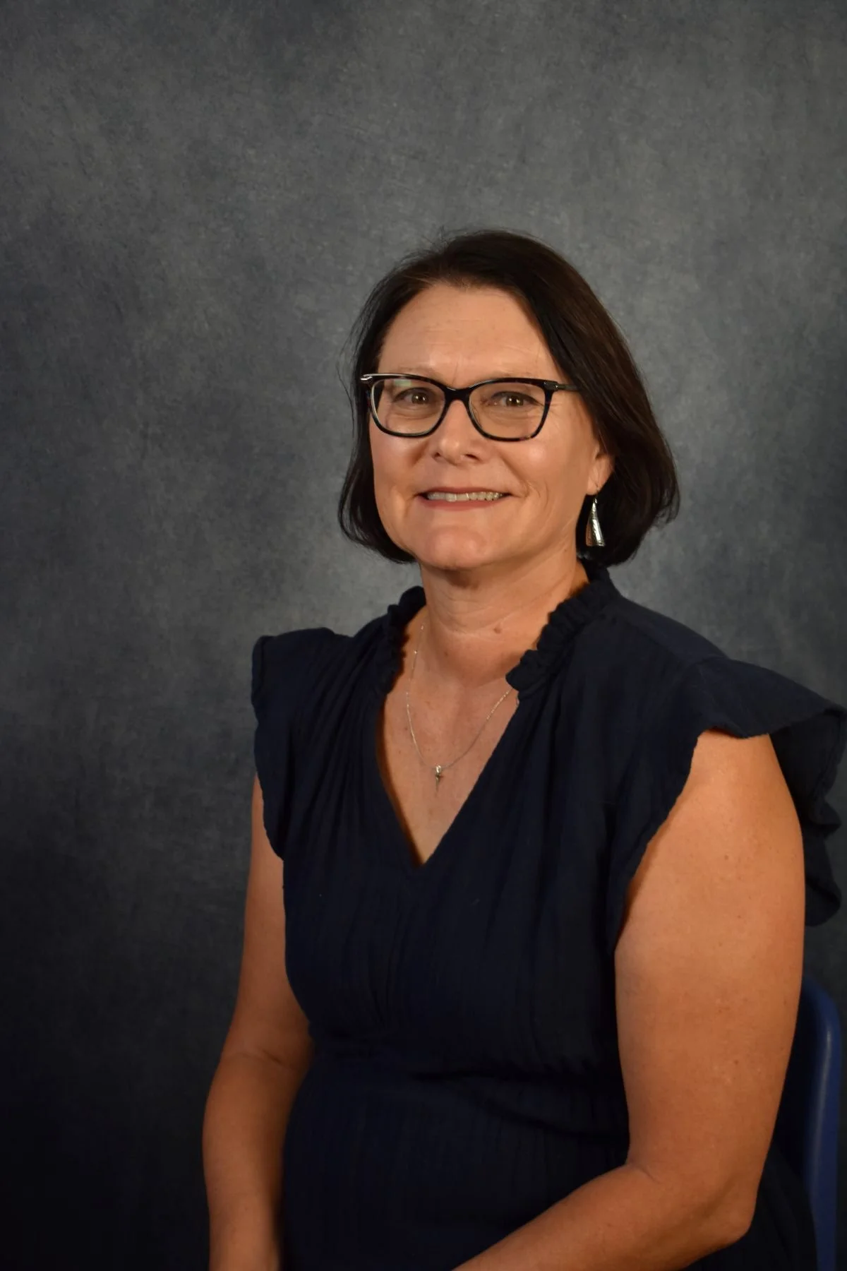 A middle-aged woman with short black hair, glasses, and earrings, smiling, wearing a dark blue ruffled sleeve top, seated against a gray background.