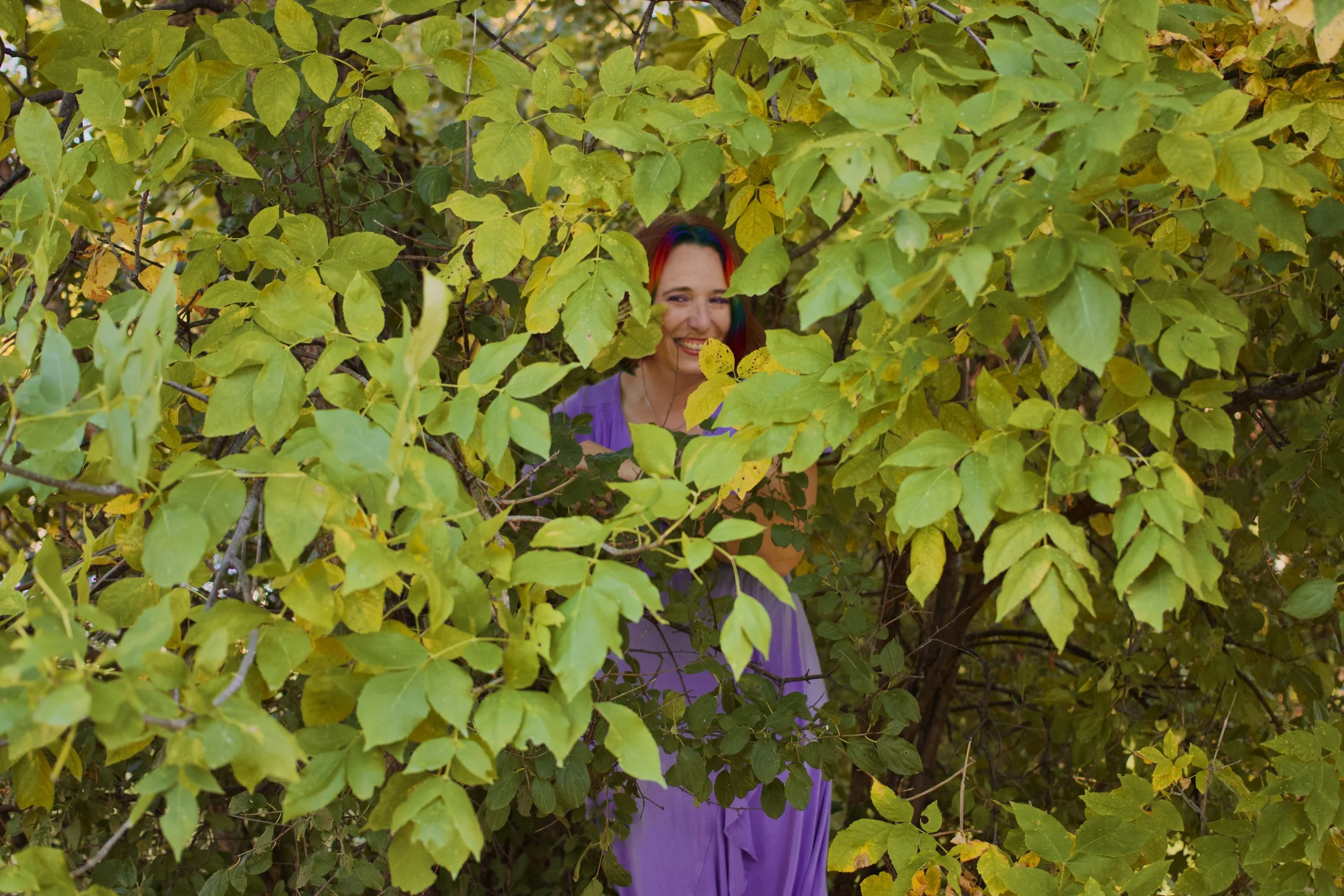 A person with rainbow-colored hair smiling through green leafy branches in a tree.