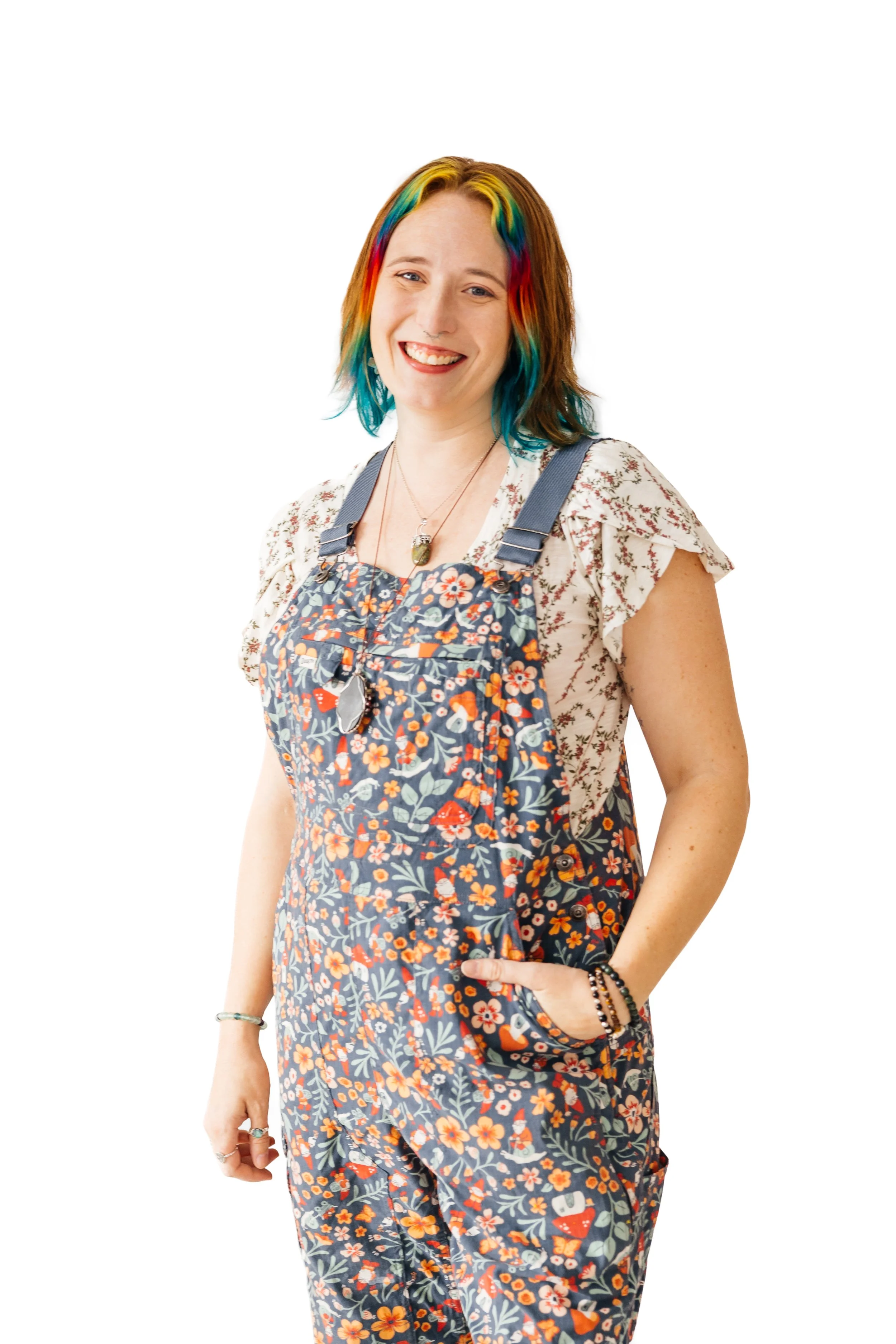 A smiling woman with rainbow-colored hair, wearing floral overalls, standing against a white background.