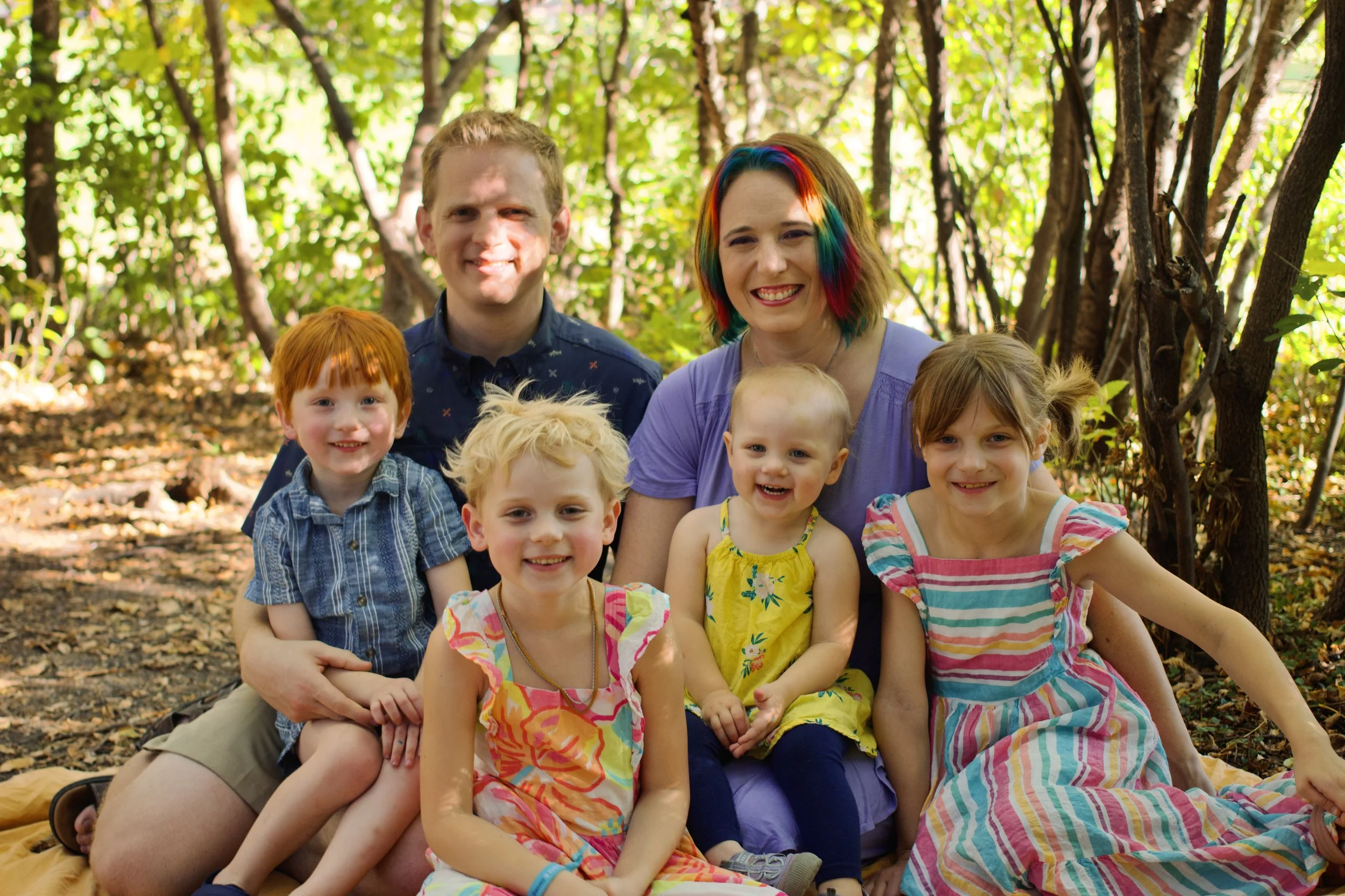 A group of children and two adults smiling in a wooded outdoor setting during autumn. The adults are sitting behind the children, who are wearing colorful summer dresses and a blue shirt. The woman has rainbow-colored hair and is wearing a purple shirt. The children have various hair colors, including red, blonde, and light brown. The background shows trees with green leaves and sunlight filtering through.