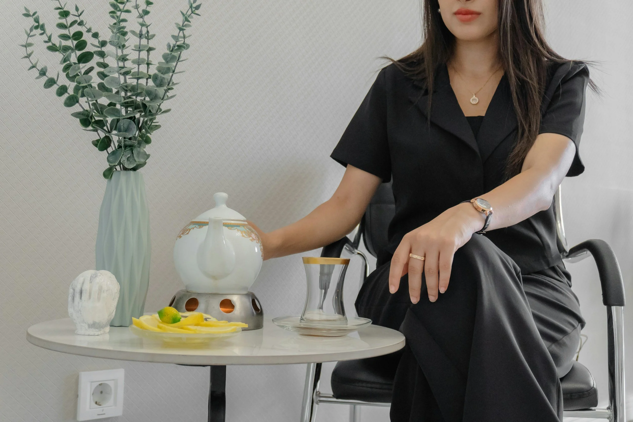 Part of a woman sitting at a table with tea and decorative items, including a tall green vase with leafy branches, a ceramic teapot, a glass teacup, and a plate with lemon slices and limes.