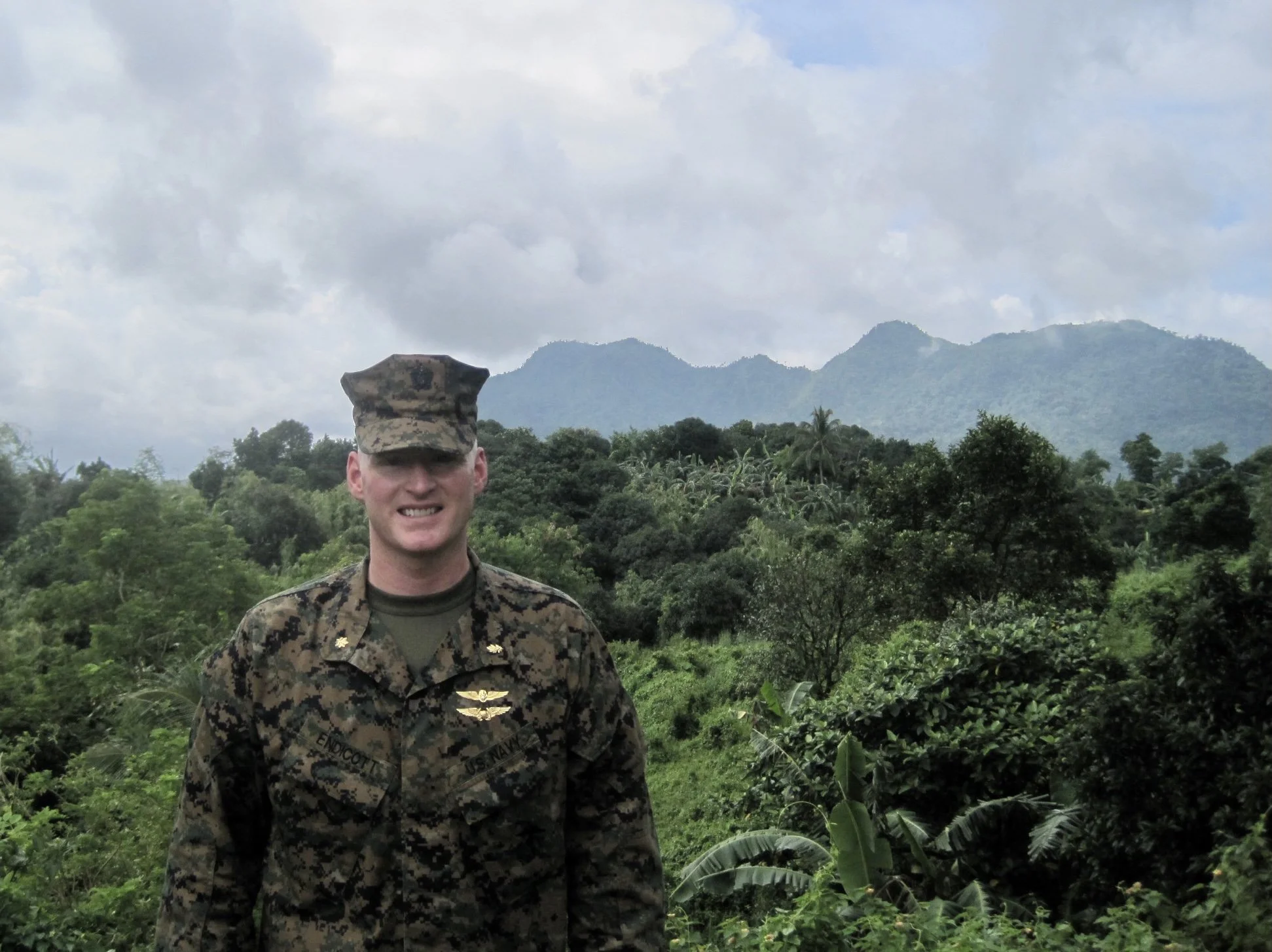 A man in military uniform standing outdoors with lush green hills and mountains in the background, under a cloudy sky.