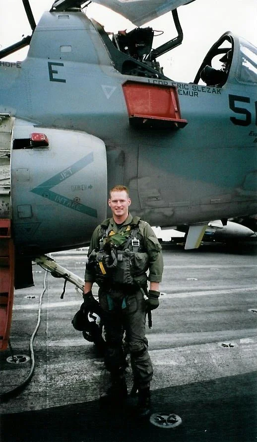 A young man in military flight gear standing on an aircraft carrier deck with a gray fighter jet in the background.
