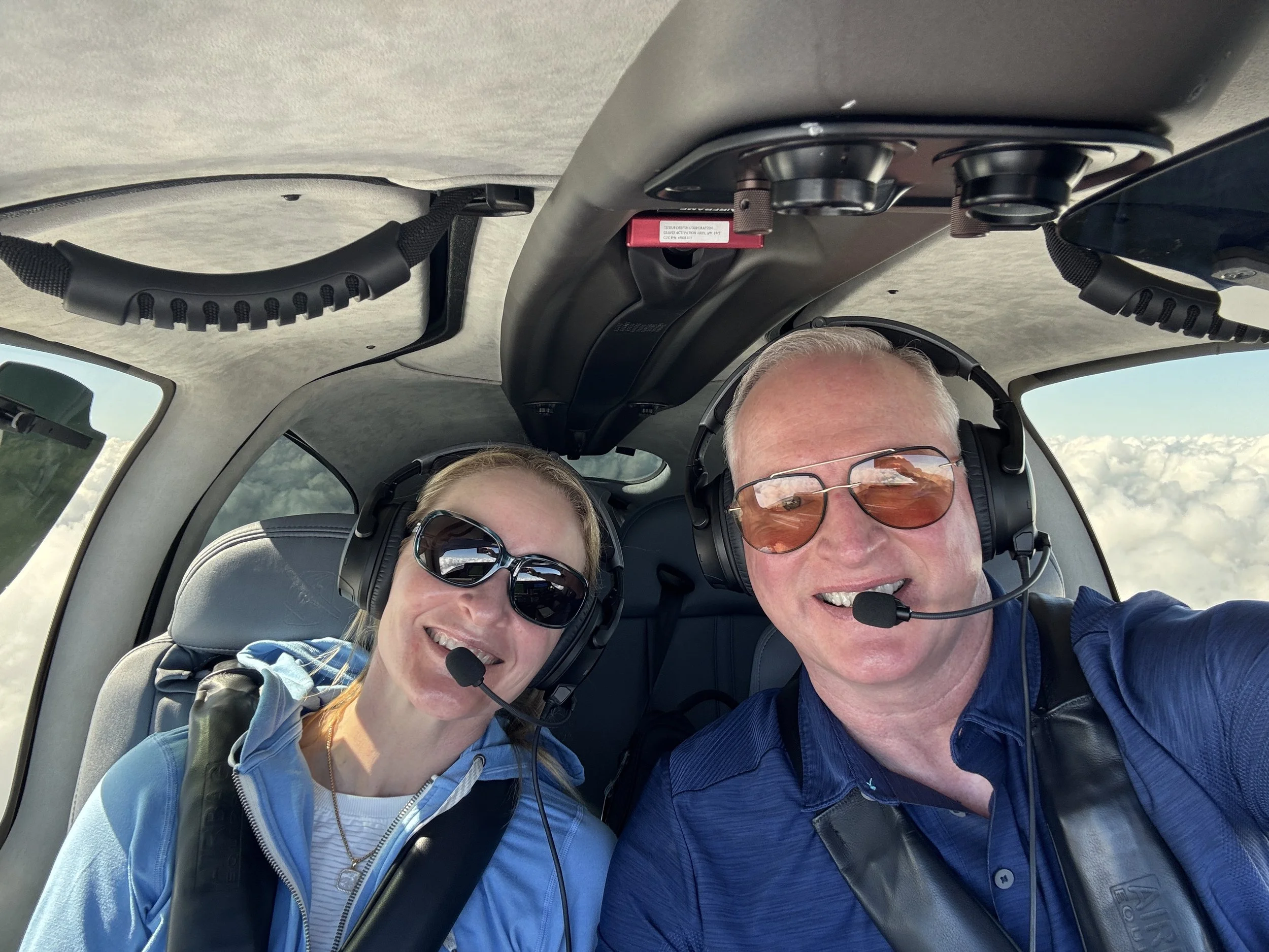 Two smiling people wearing headsets and sunglasses, sitting inside a helicopter, with cloudy sky visible through the windows.