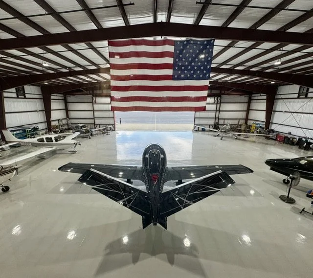 Interior of a large airplane hangar with several small aircraft, an American flag hanging from the ceiling, and a sleek black private jet in the center.