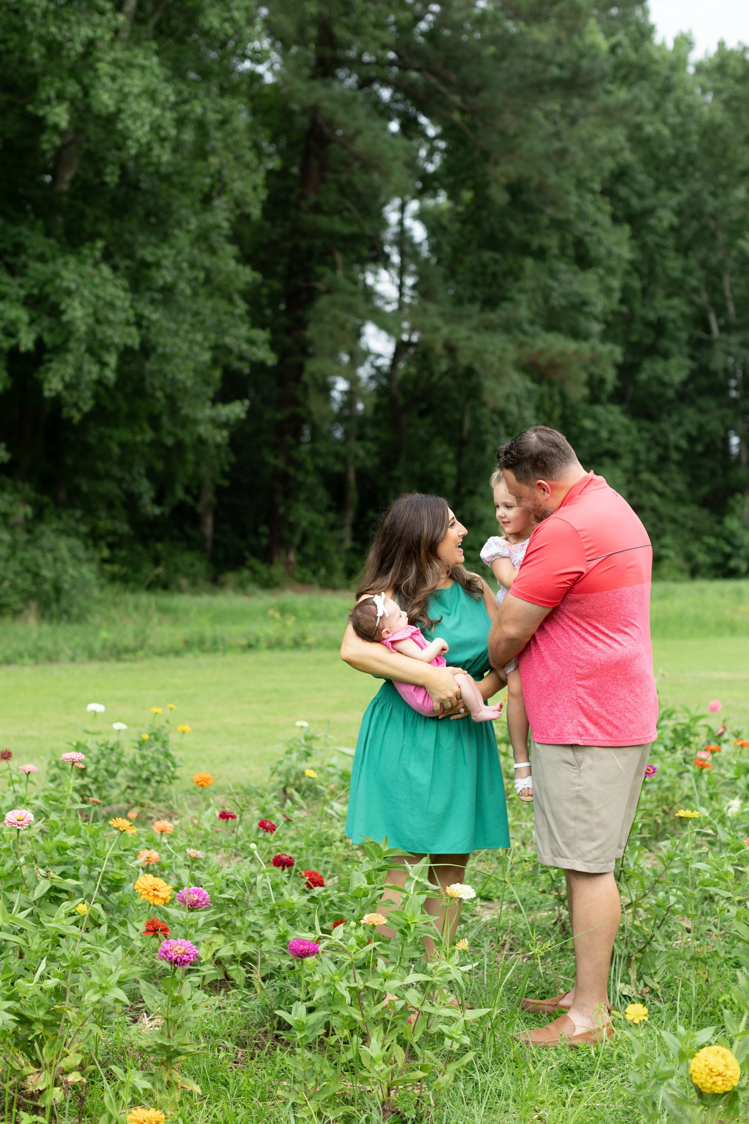 A family of four, including two women and two children, standing in a flower garden with trees in the background. They are smiling and looking at each other.