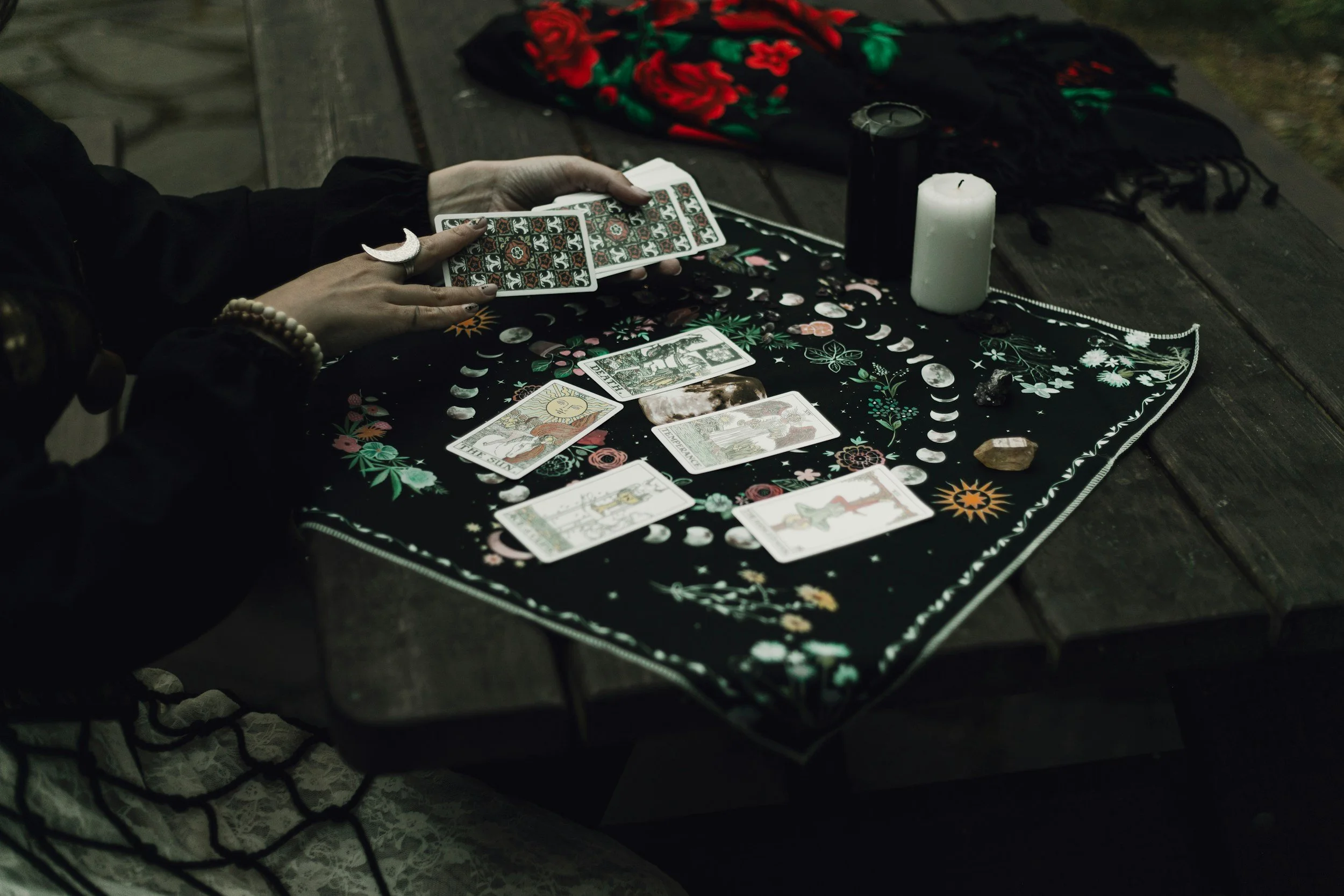 A person holding tarot cards over a decorated cloth, with candles and stones on a wooden surface, surrounded by a patterned scarf and a red-flowered fabric.