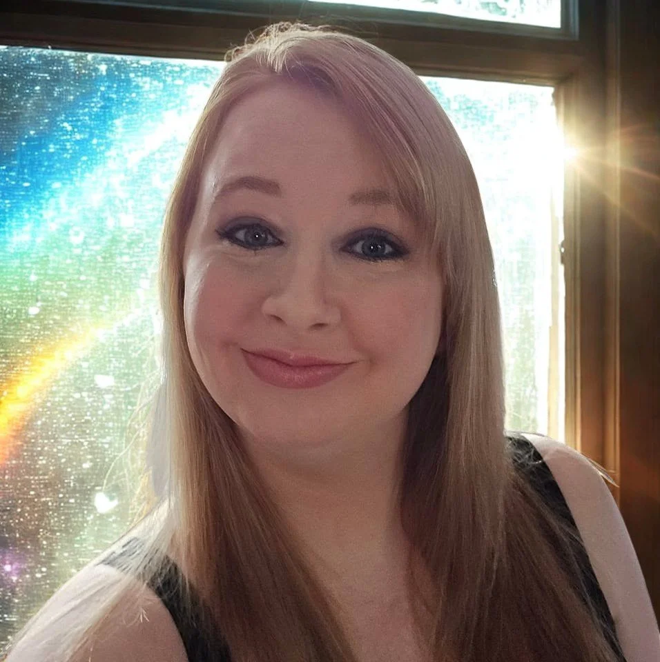 A smiling woman with long red hair and blue eyes standing near a window with sunlight streaming in, next to a colorful sparkly background.