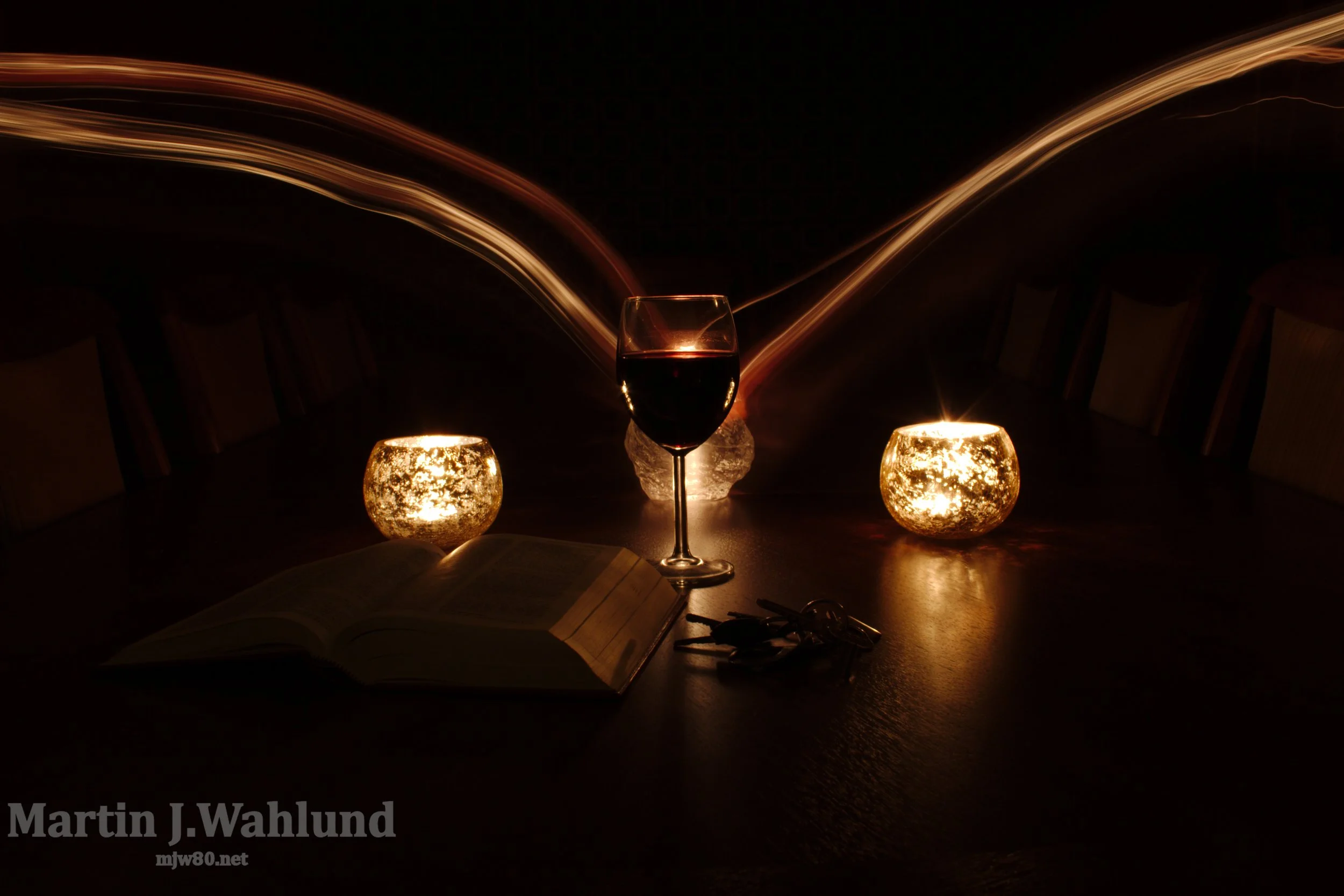 A dark table setting with a glass of red wine, two candle holders with lit candles, and an open book, with light streaks in the background.