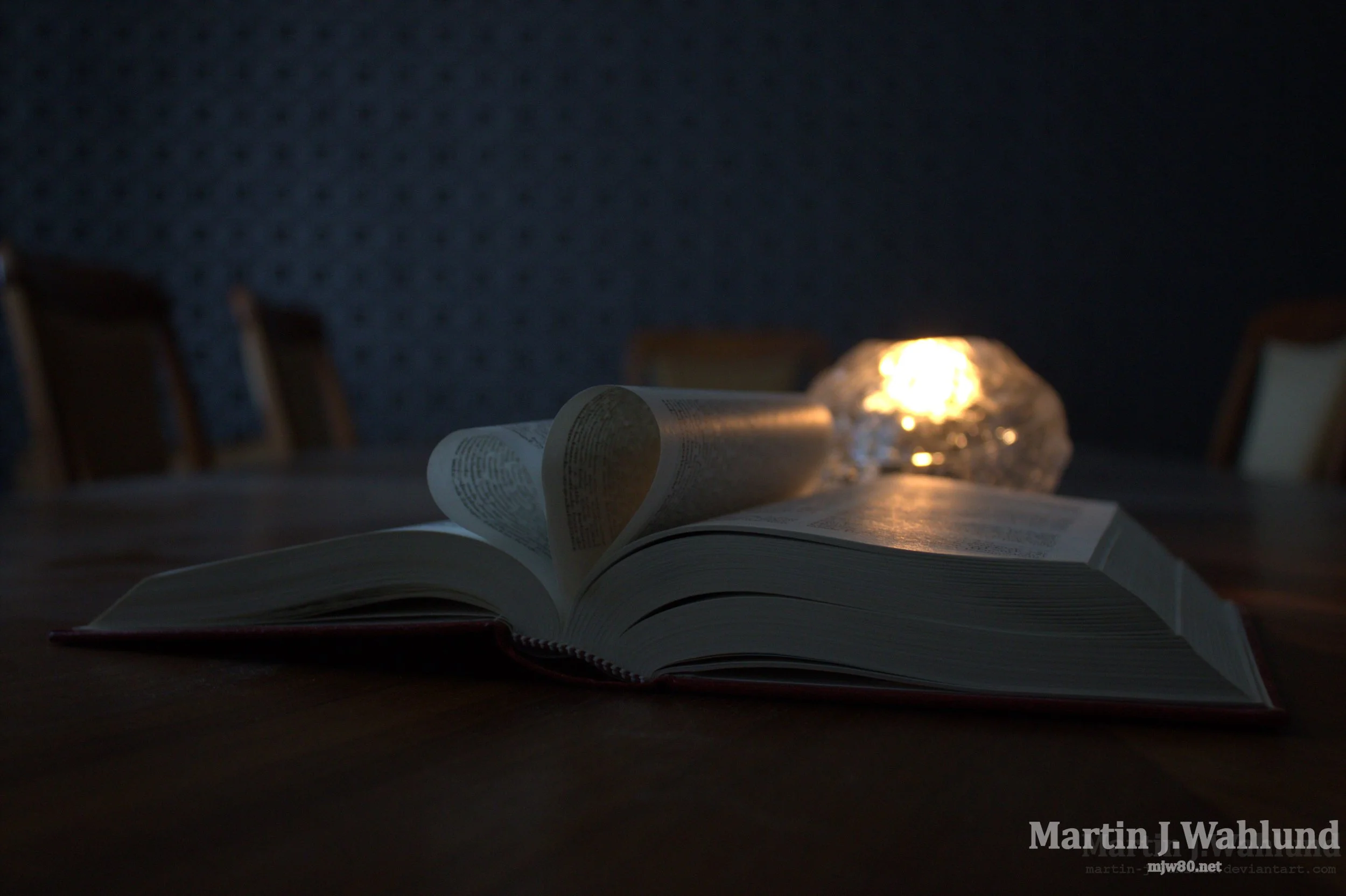 An open book on a table with a glow in the background, possibly from a candle or lamp, and several chairs around the table.