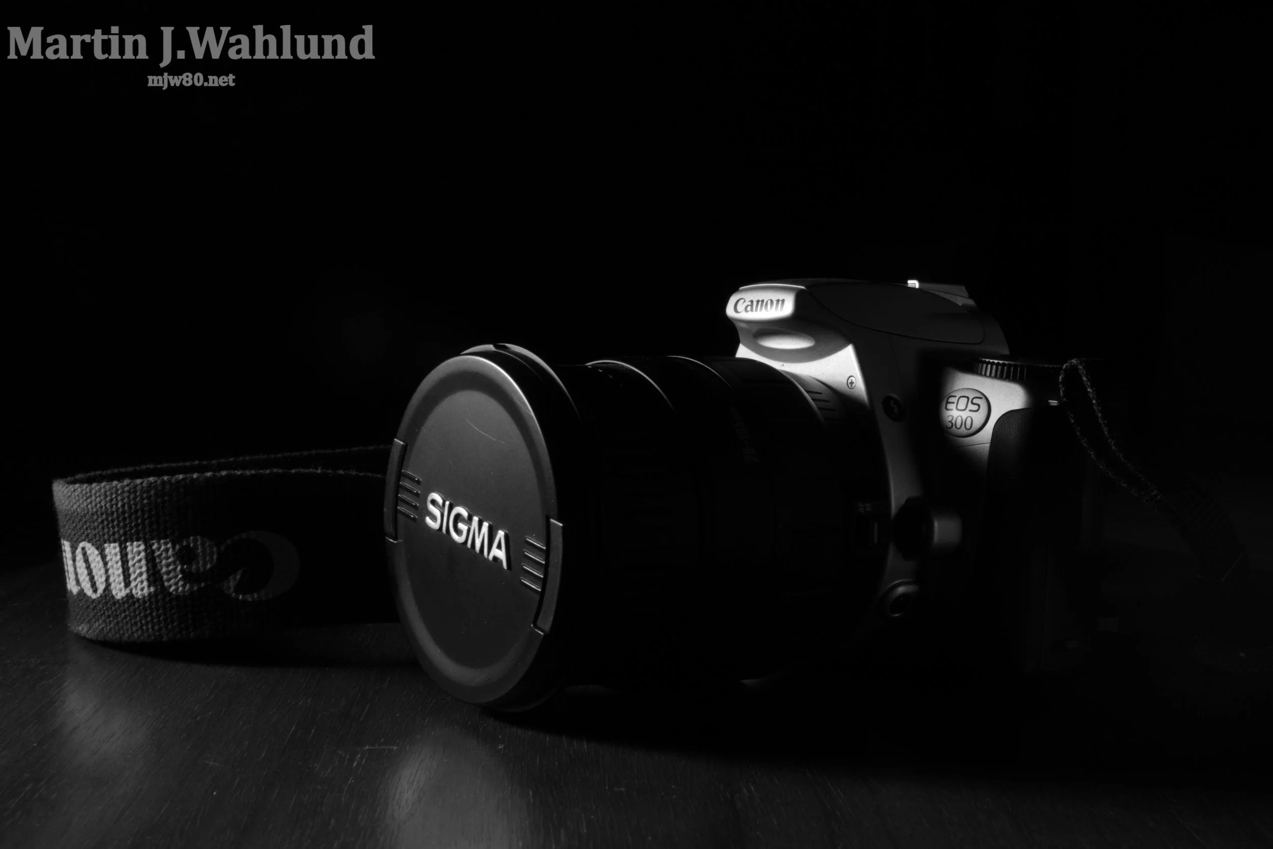 A Canon EOS 300 camera placed on a black surface with a Sigma lens attached. In the background, there is a black strap and a fabric item with the word 'LOVES' visible.