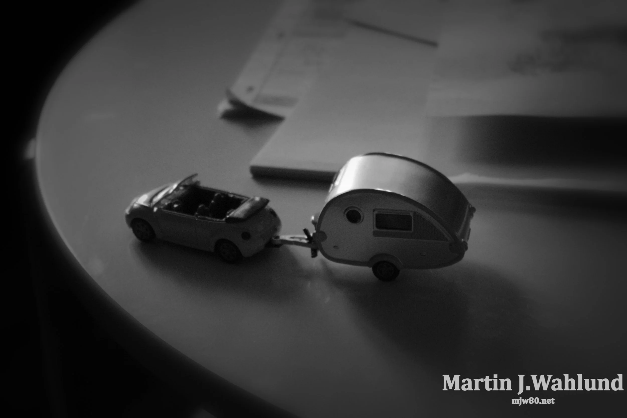 Black and white close-up view of toy cars, a camper trailer, and a small open convertible car on a plain surface, with a shadowy circular vignette border and a text watermark reading 'Martin J. Wahlund' and 'mjw80.net' in the bottom right corner.