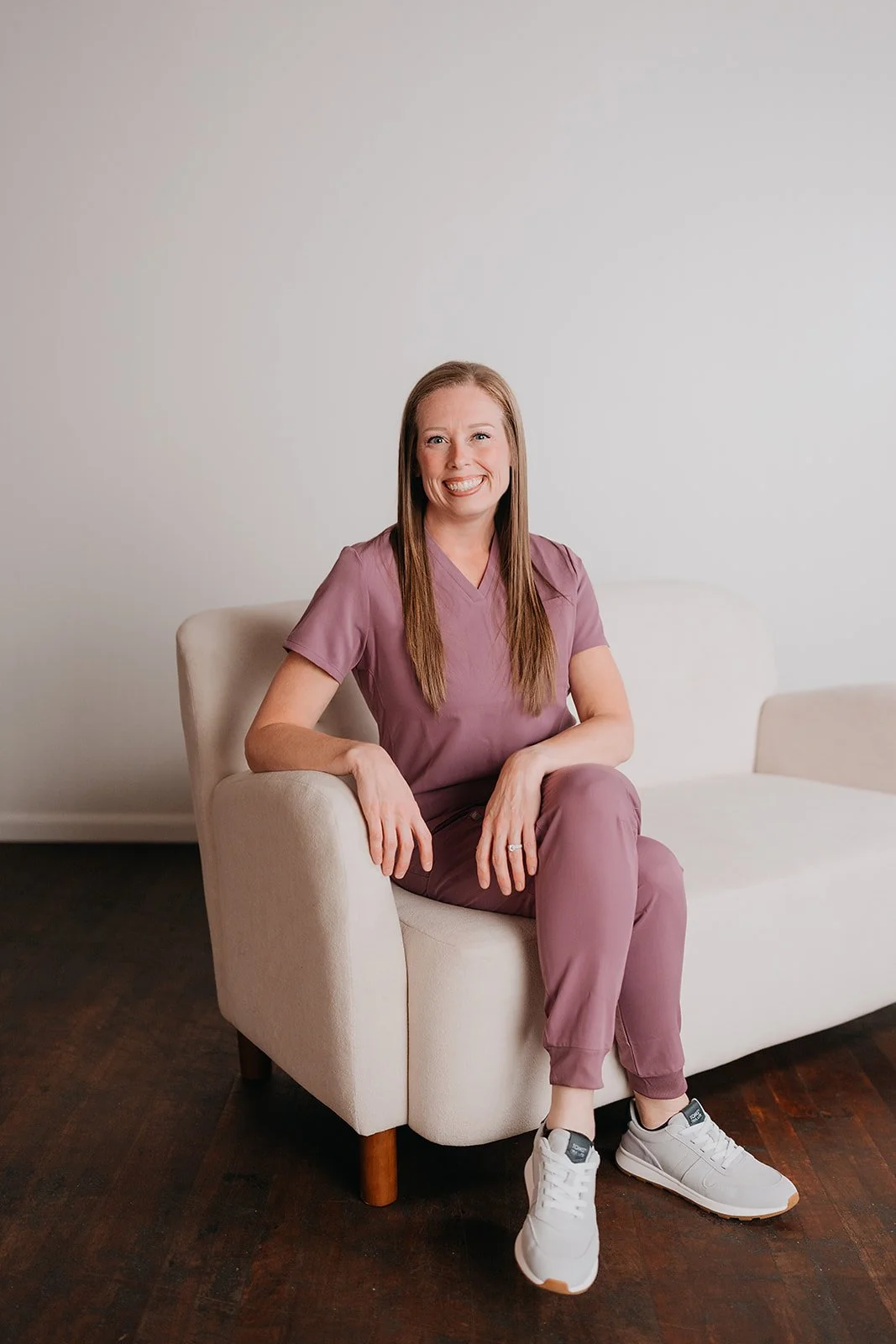 A woman with long brown hair sitting on a beige sofa in front of a plain white wall, wearing a mauve medical scrub uniform and white sneakers, smiling at the camera.