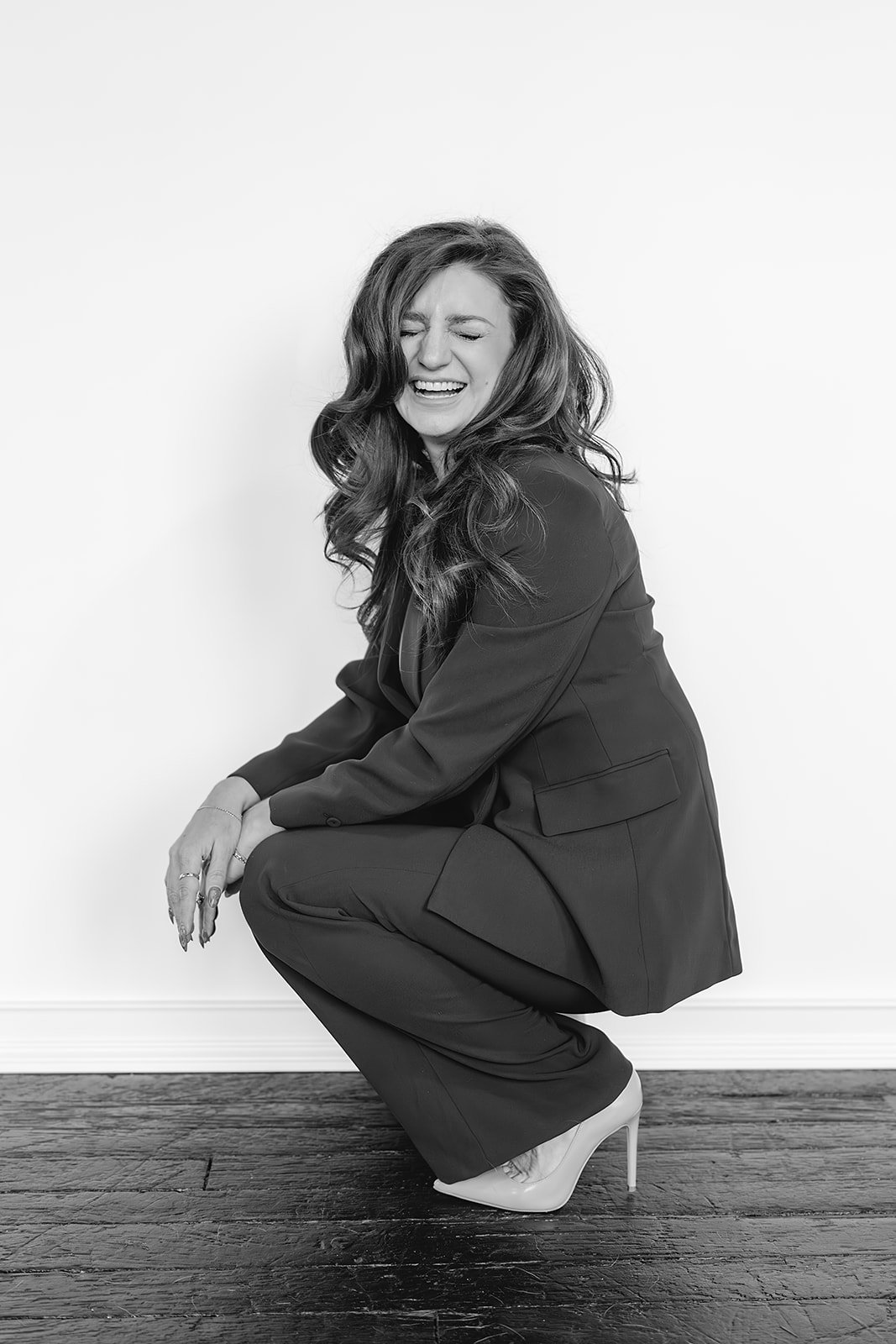 A woman with long wavy hair wearing a suit and high heels, squatting and smiling with eyes closed against a plain white wall.