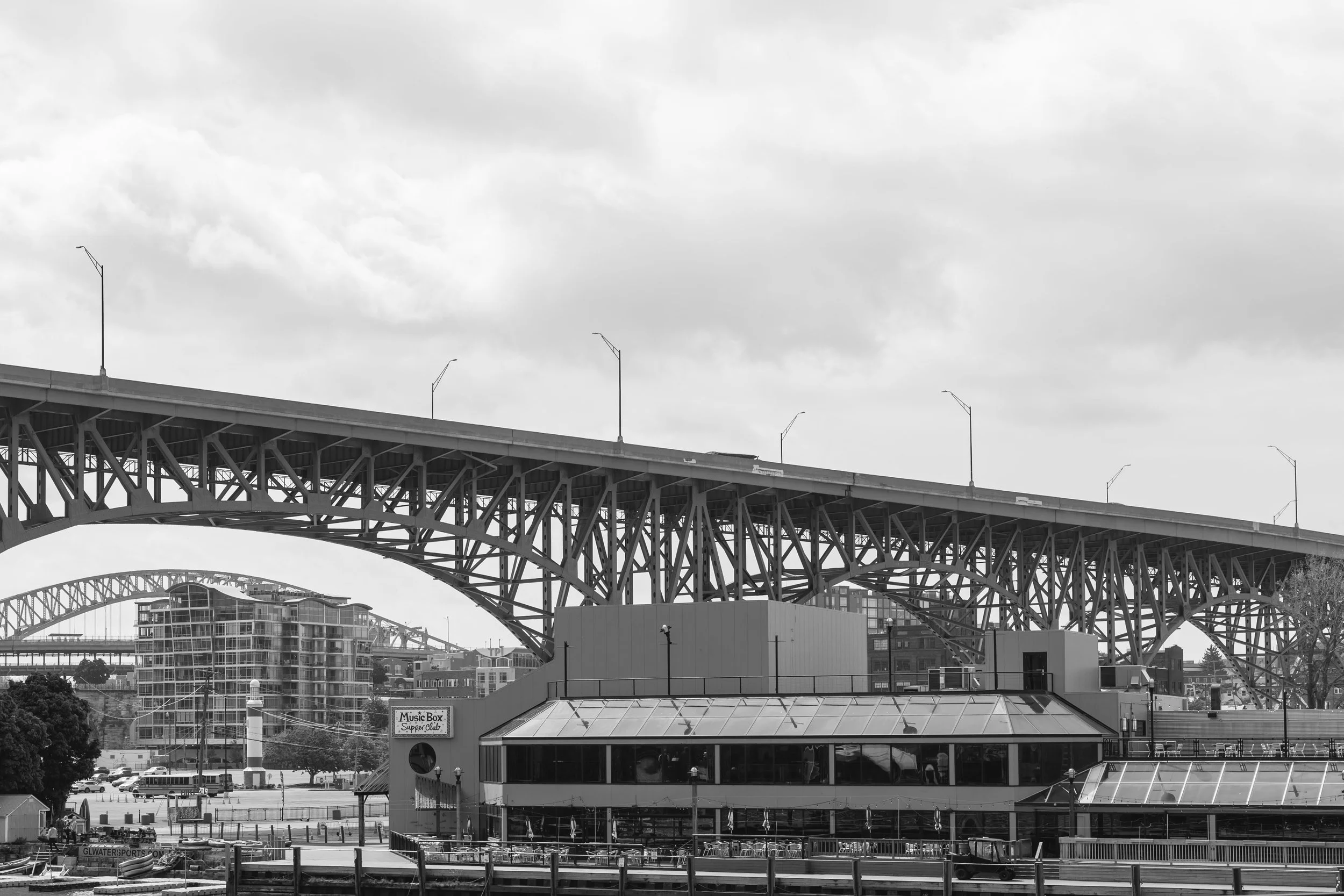 Black and white photo of a large bridge with multiple arches and streetlights, over an urban area with buildings, trees, and a waterfront with boats.