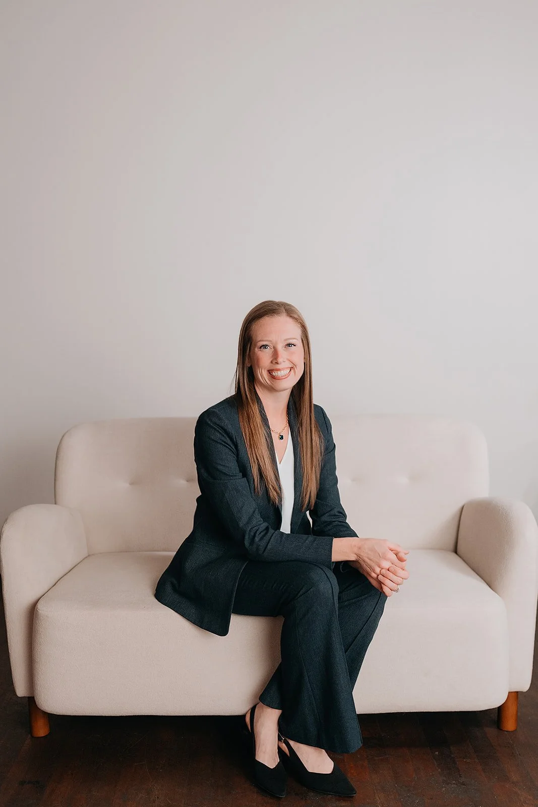 A woman with long red hair, wearing a dark blazer and pants, white top, and black heels, sitting on a beige sofa smiling at the camera.
