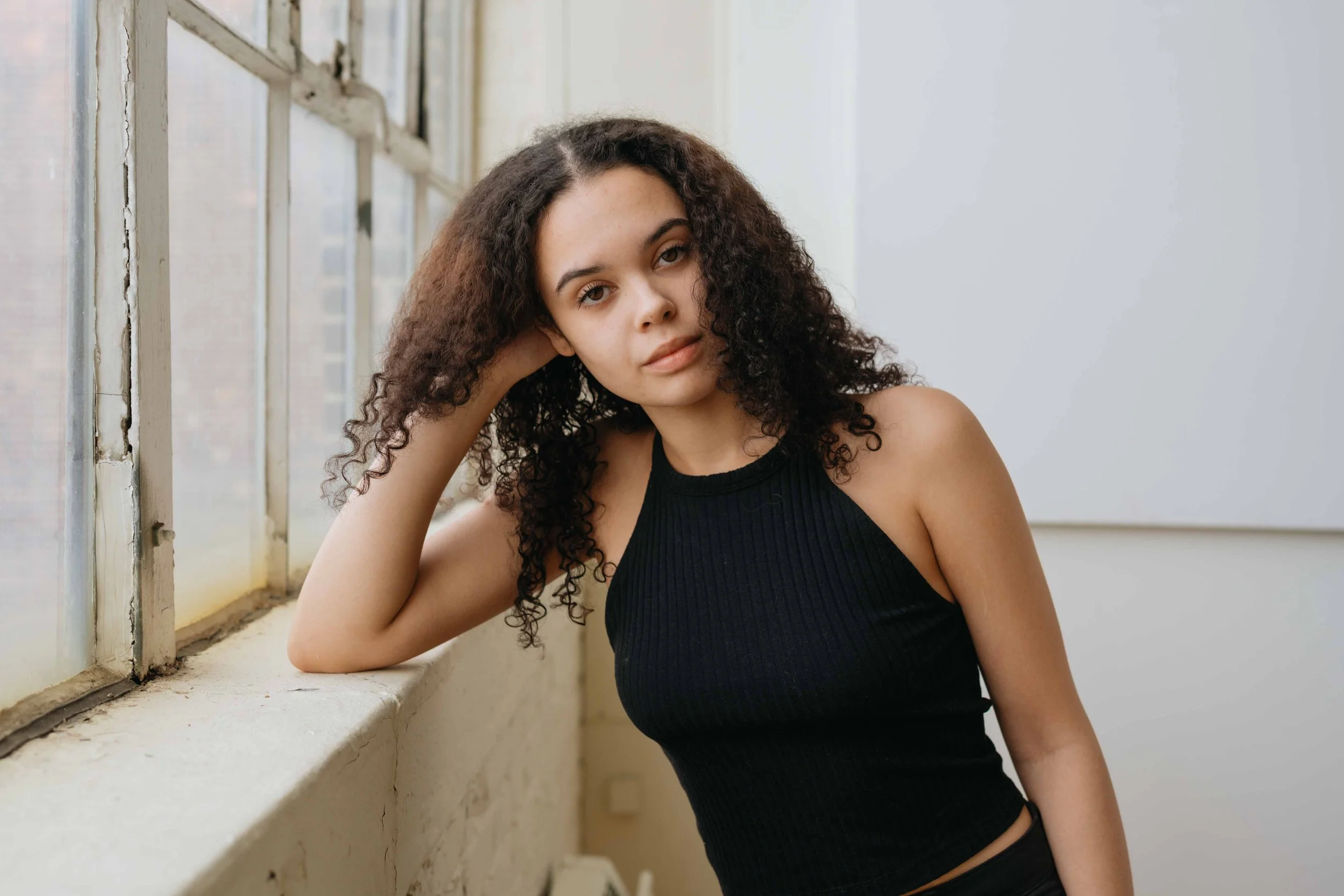 A young woman with curly brown hair sitting by a large window with a white frame, resting her head on her hand, wearing a black sleeveless top, looking at the camera.