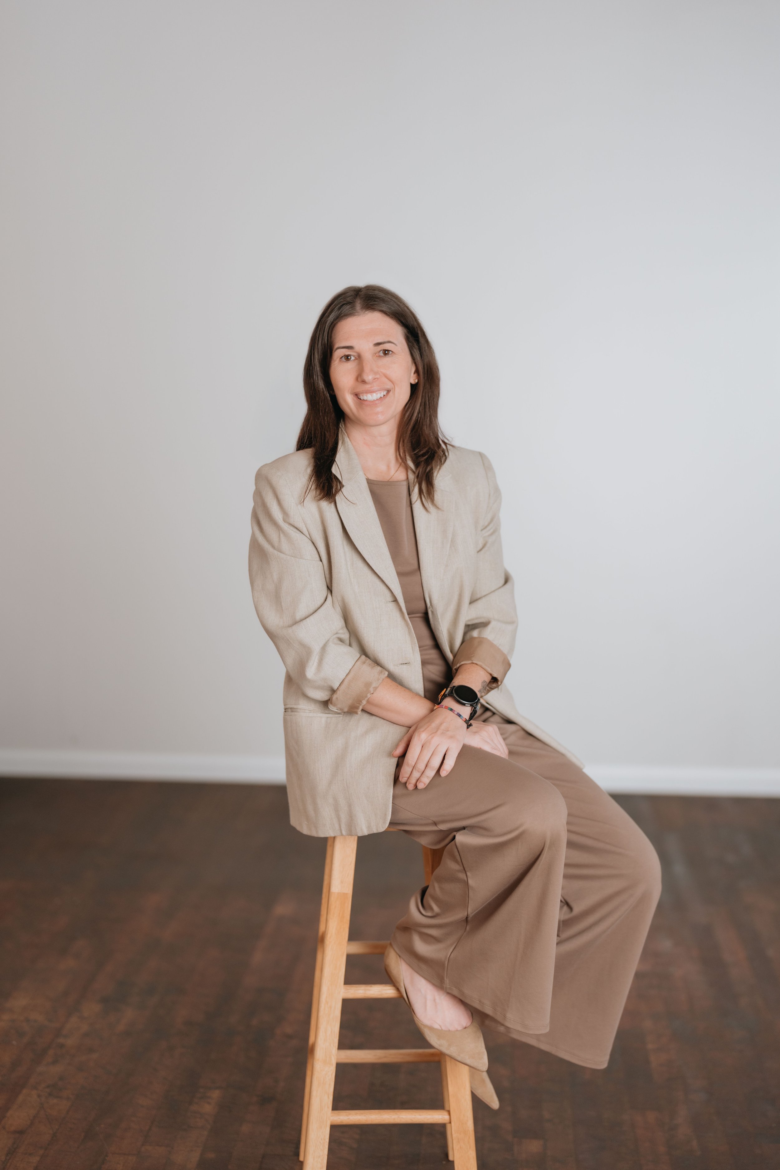 A woman with long brown hair, wearing a beige blazer and brown pants, sitting on a wooden stool in front of a plain white wall and brown hardwood floor, smiling at the camera.