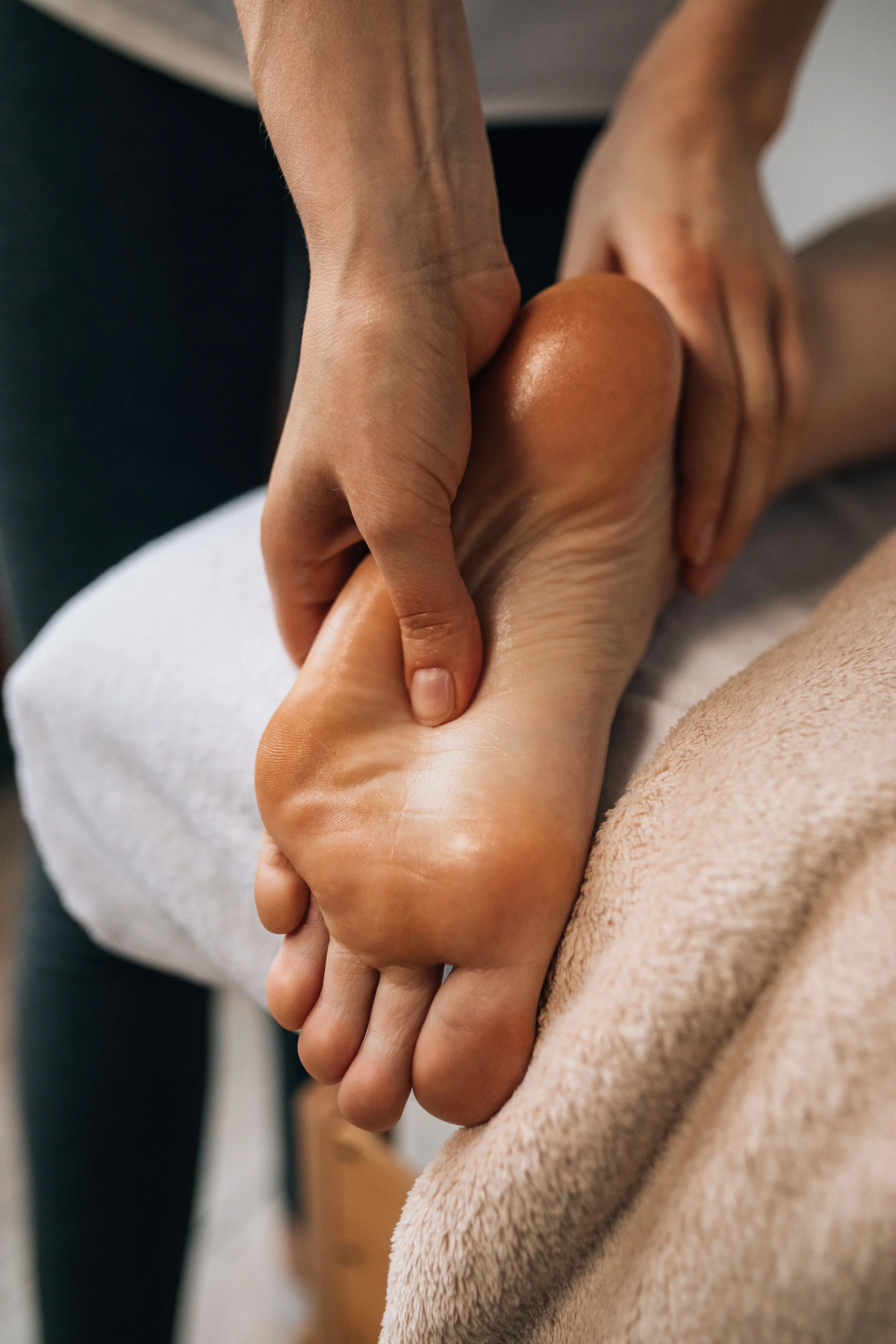 A close-up of a person's hand with slender fingers holding the heel of another person's foot, along with a soft pink blanket or towel partially covering the leg, suggesting a foot massage or care setting.