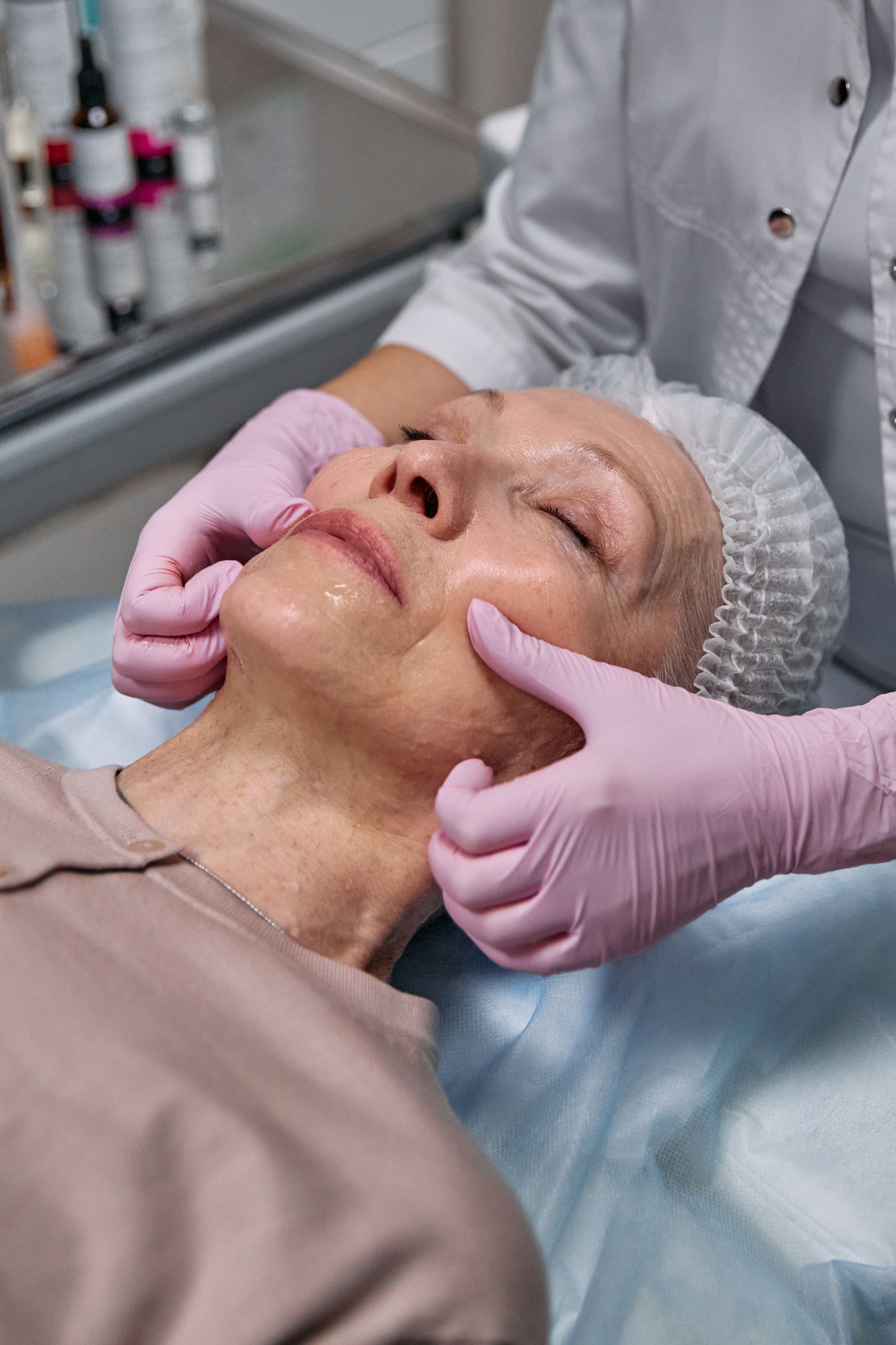 A woman receiving a facial treatment at a spa. She is lying down with her eyes closed, wearing a hair cap and a beige top. A person wearing pink gloves is gently pressing on her face. In the background, there are bottles and skincare products on a tray.