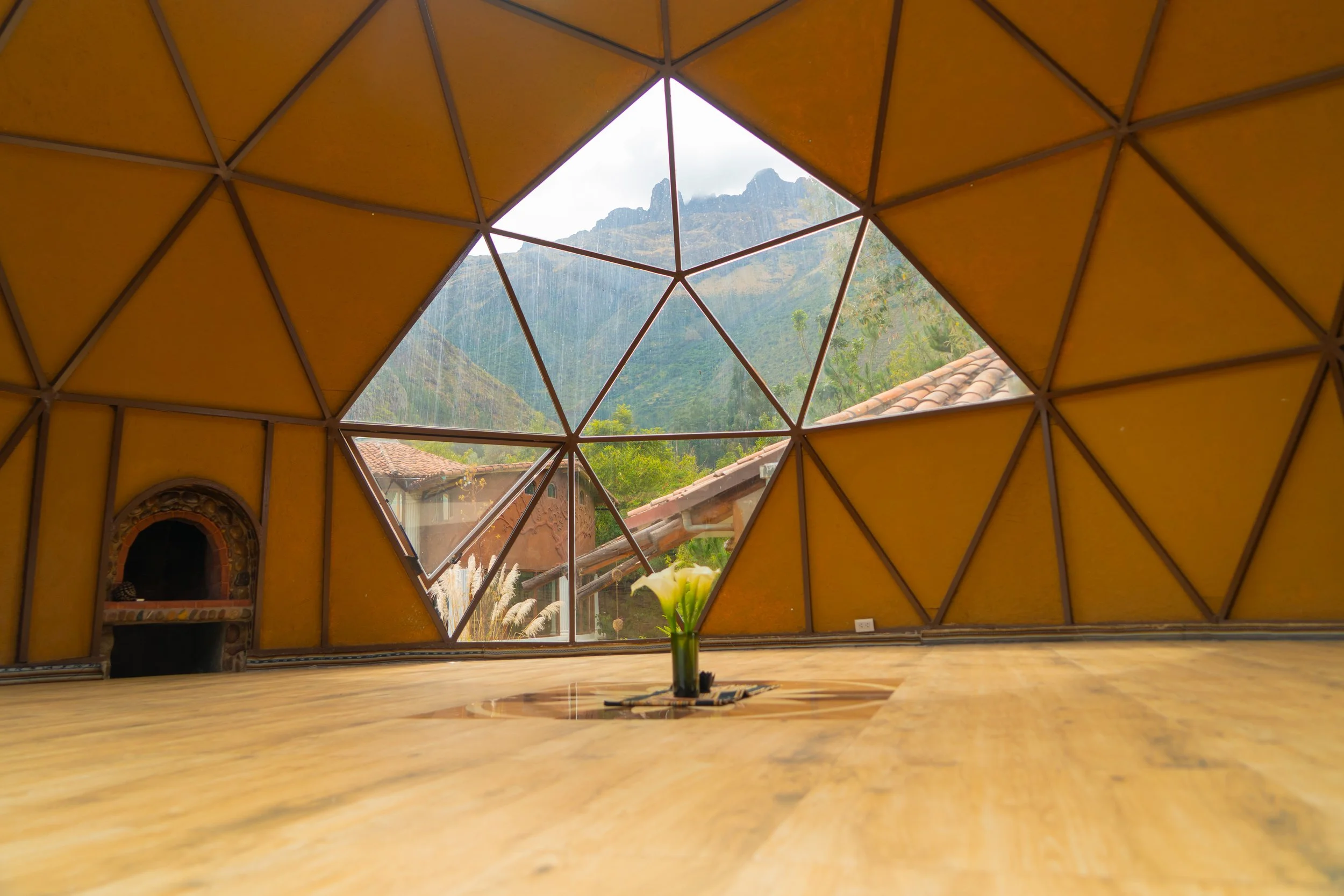 Interior of a dome-shaped wooden structure with geometric patterns, view through a large polygonal window showing mountains, trees, and rooftops, with a vase of white flowers on the wooden floor.