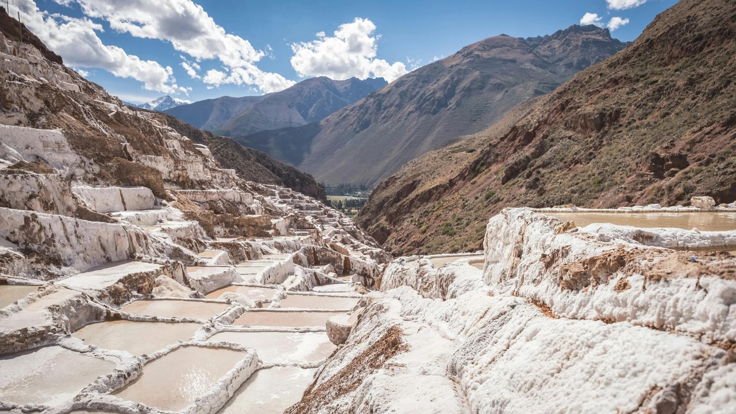 Terraced salt flats set in mountainous terrain with brown and green slopes, under a partly cloudy sky.