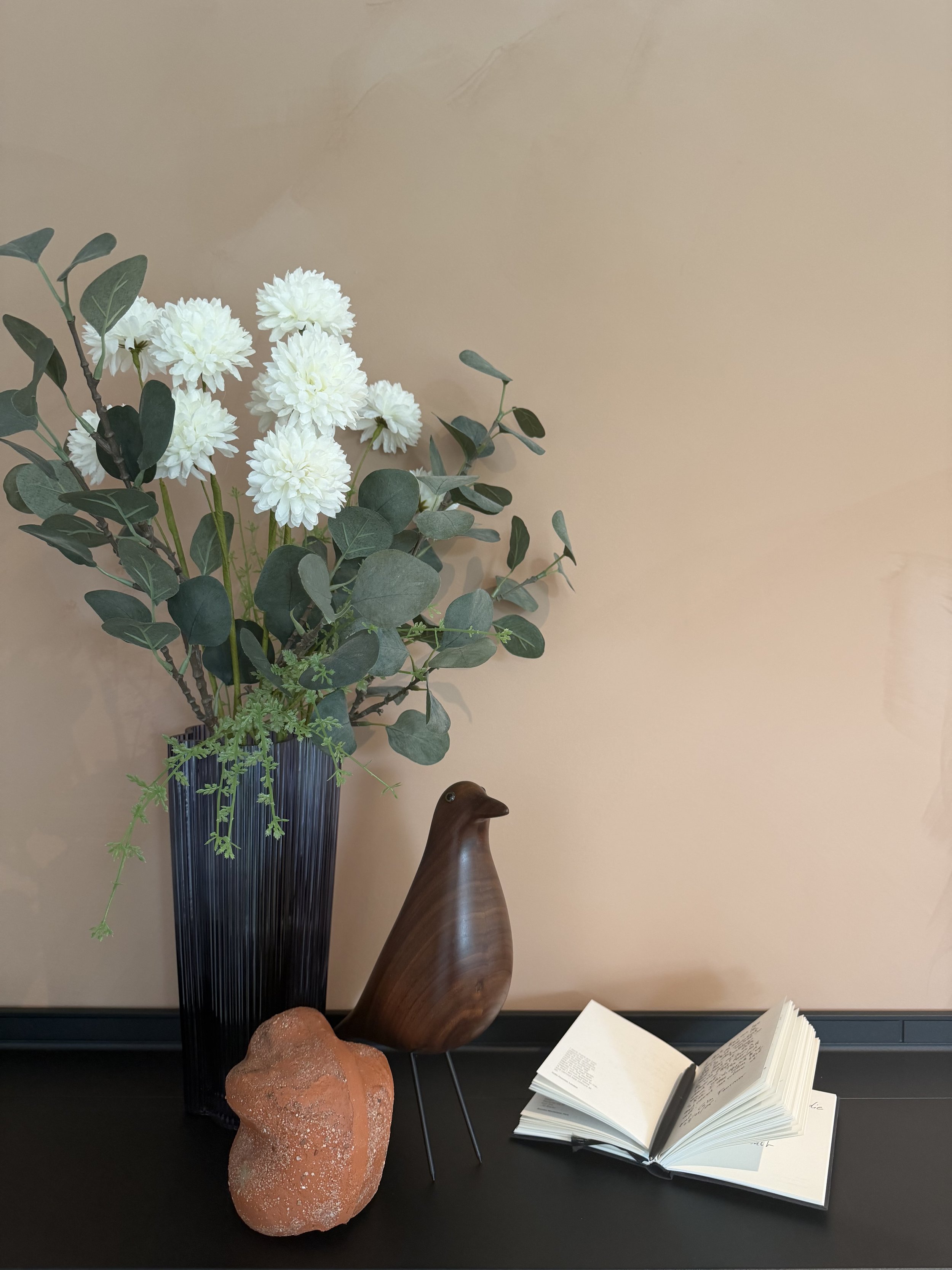 Een vaas met witte bloemen en groene bladeren, een houten vogelbeeld, een rots, een open boek met een pen, alles op een zwarte tafel voor een beige muur.