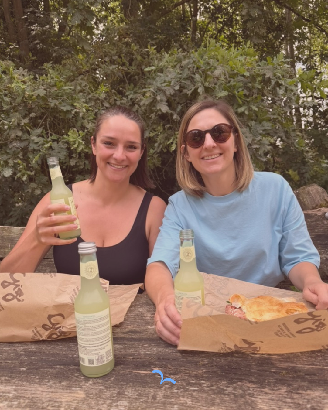 Twee vrouwen zitten buiten aan een houten tafel met pizza en frisdrank in een groene, bosrijke omgeving.