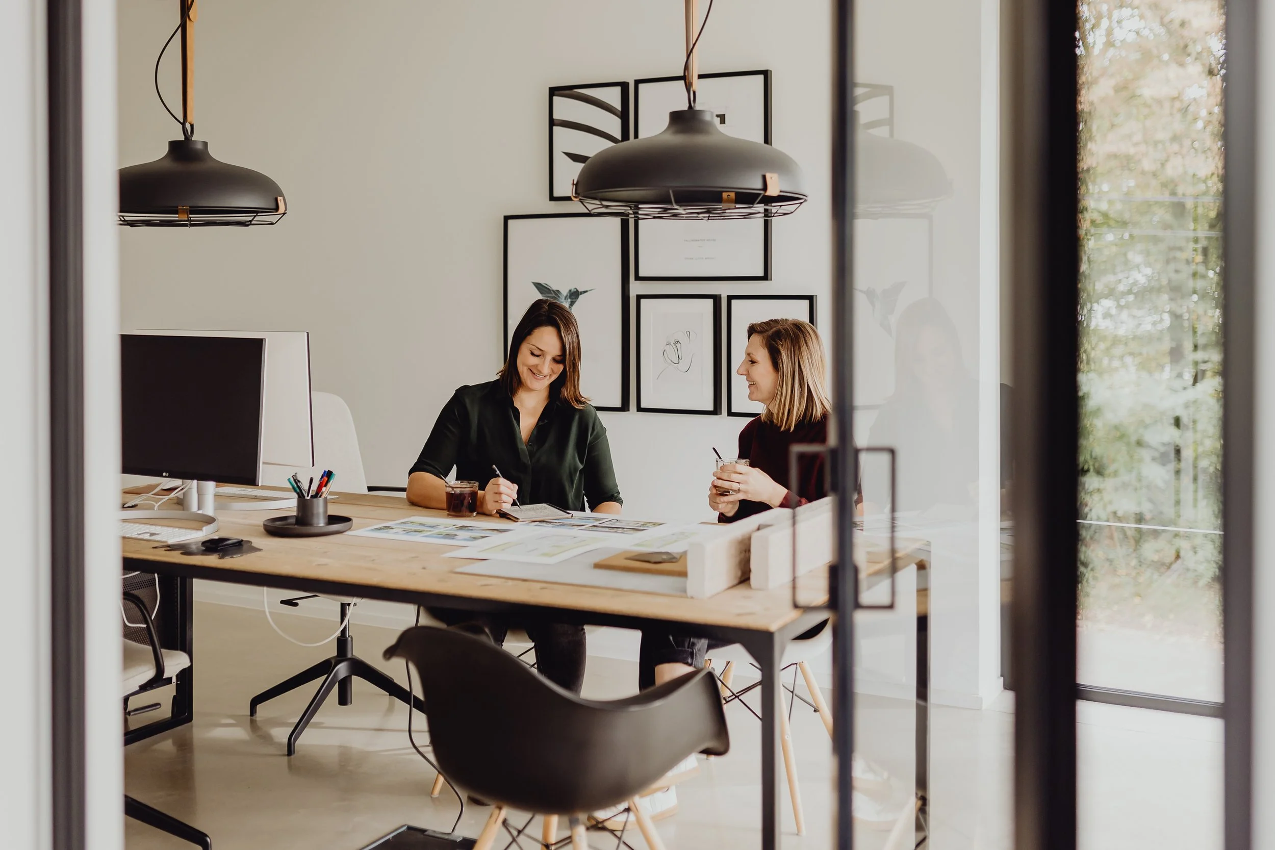 Twee vrouwen die binnen aan een bureau zitten, lachend en met drinken in hun handen, terwijl ze samenwerken in een modern kantoor met kunstwerk aan de muur.
