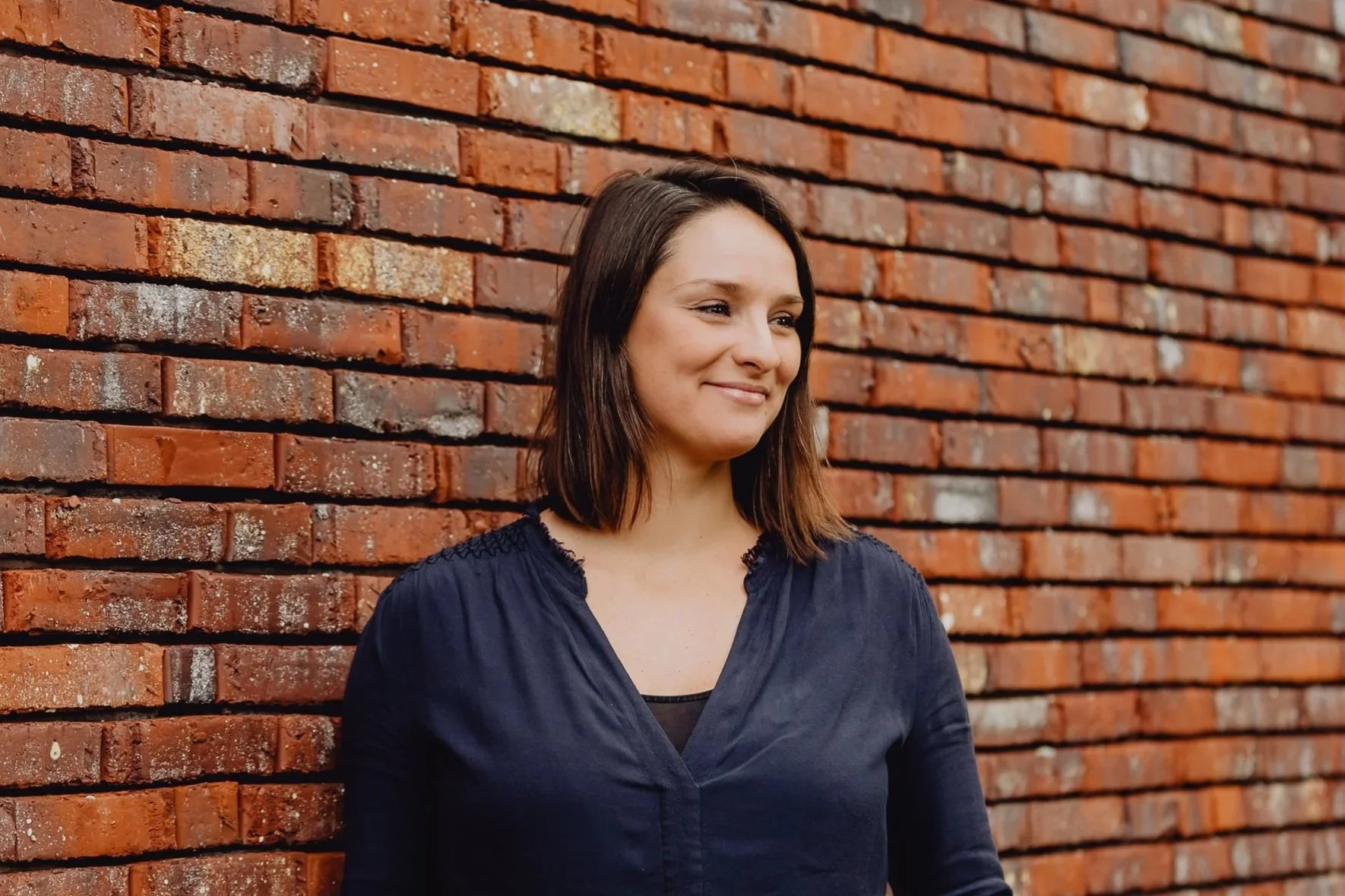Een vrouw met bruin haar die glimlacht, staande voor een rode bakstenen muur.