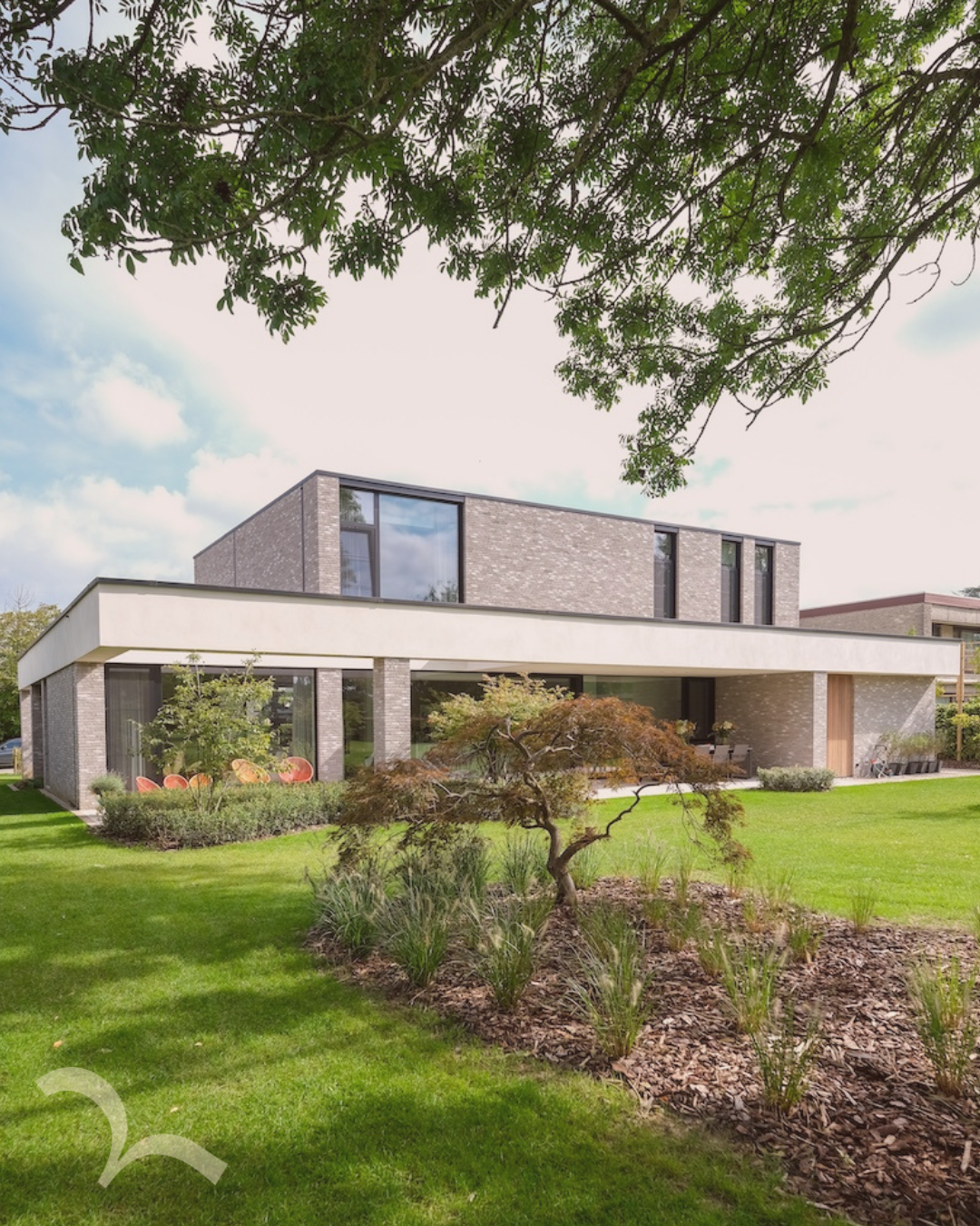 Moderne woning met grote glasramen, een tuin met groene gras, een kleine boom, en kleurrijke stoelen onder een boom, met een lichtblauwe hemel en wat wolken.