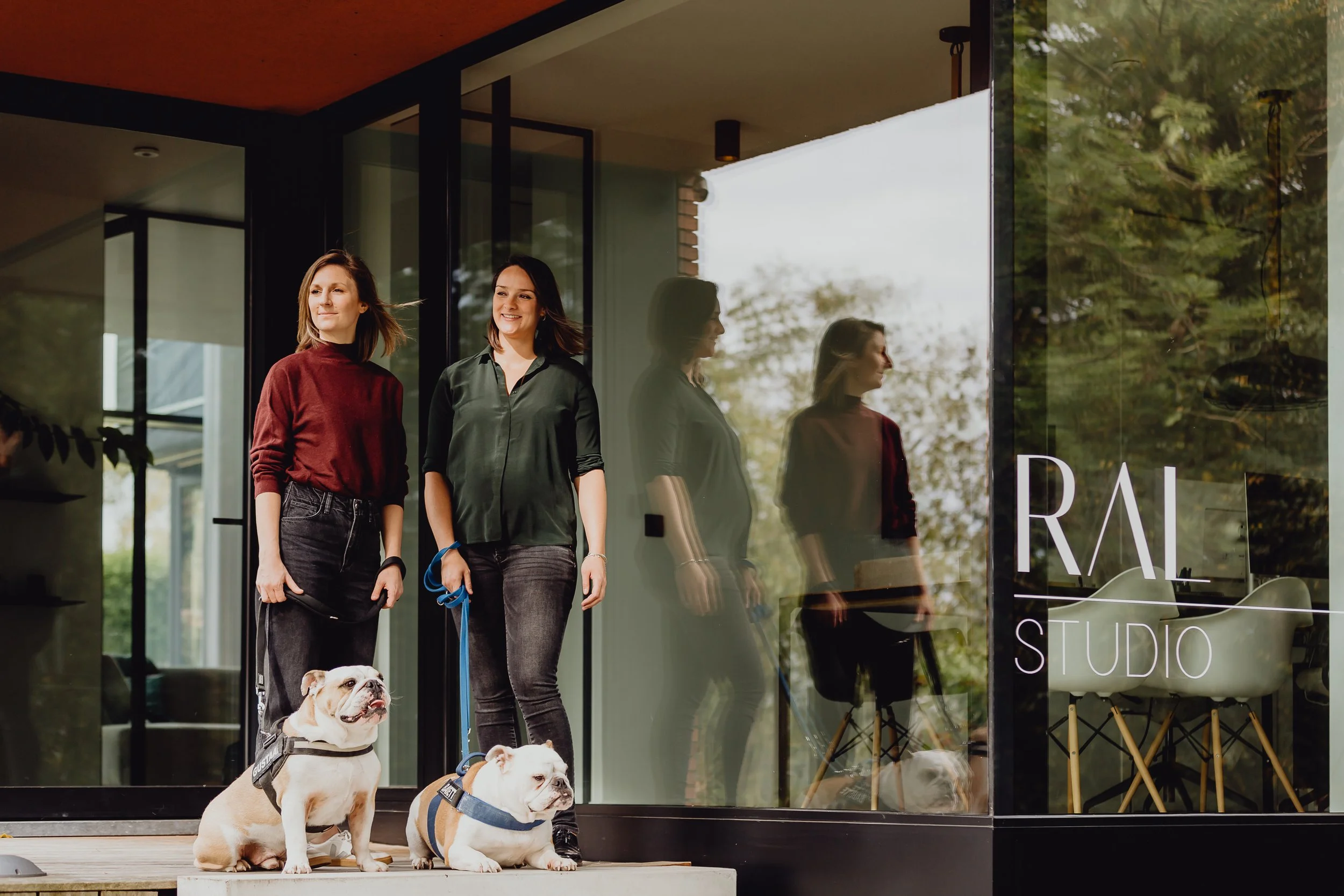 Twee vrouwen staan voor een modern gebouw met grote ramen. Ze glimlachen en houden honden aan de lijn. Een hond zit op de grond, een andere ligt. De reflectie in het raam toont meer mensen en een boomgaard.