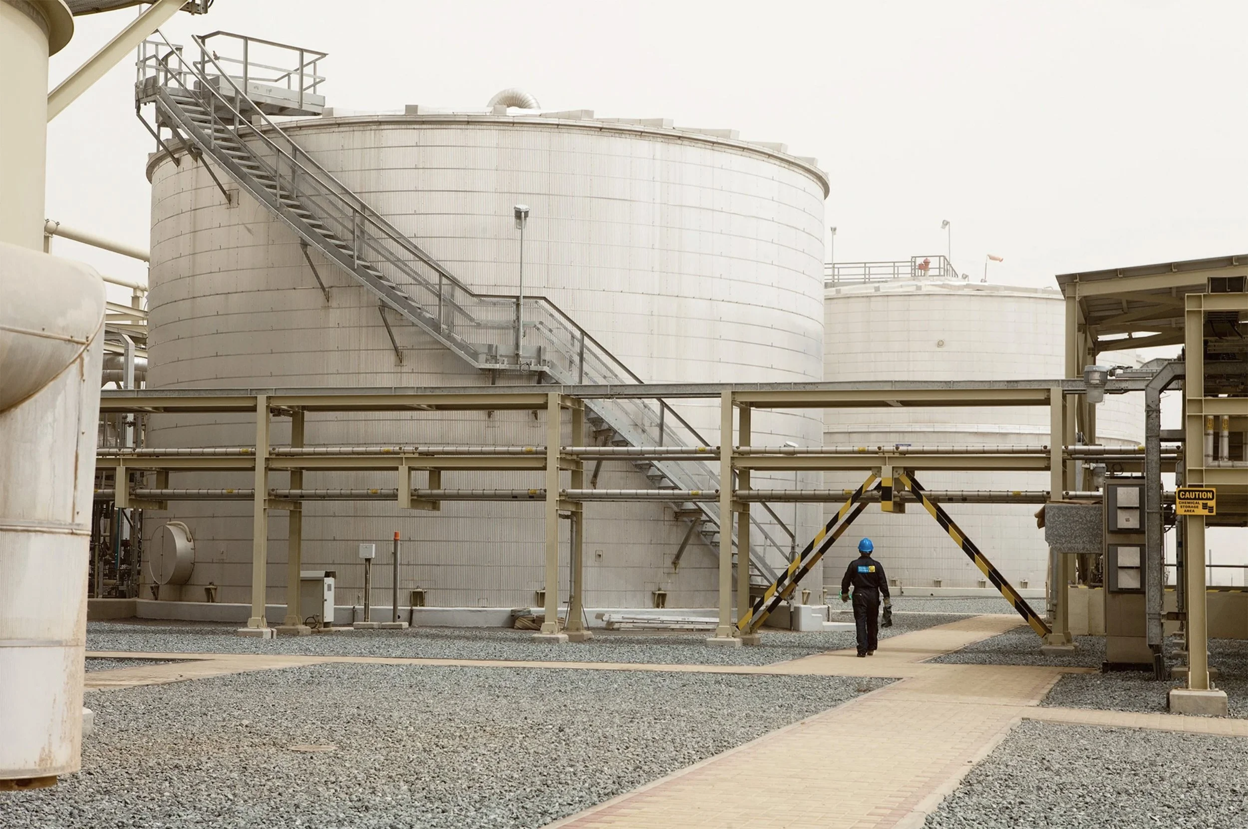 Chevron worker walking through an industrial facility with storage tanks and pipelines.