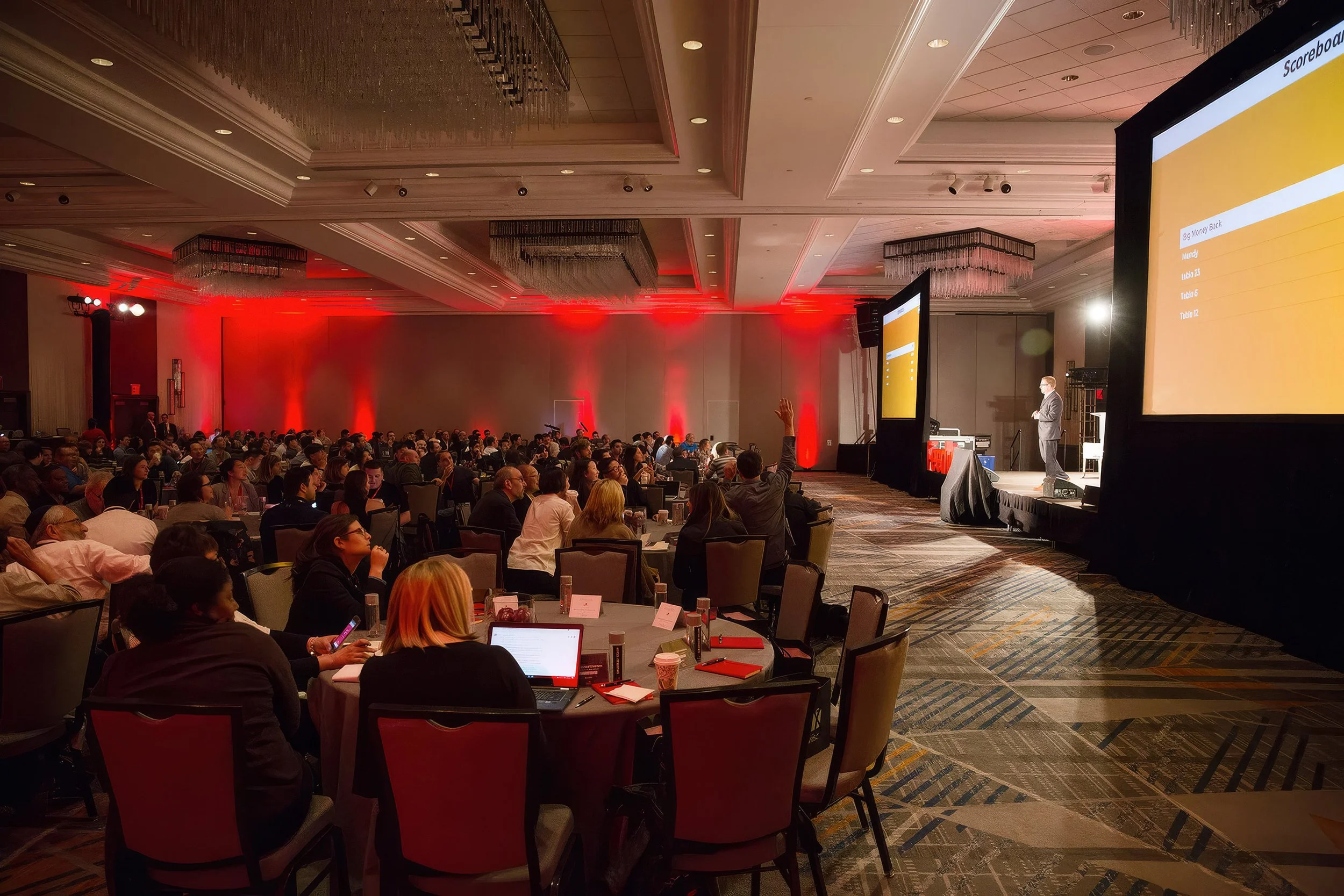 Corporate conference audience watching a presenter on stage in a red-lit ballroom.