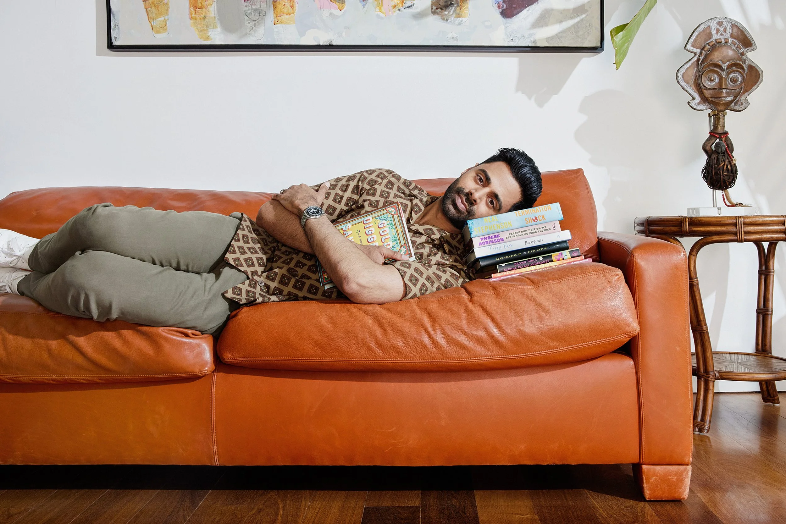 Hasan Minhaj reclining on an orange sofa with a stack of books.