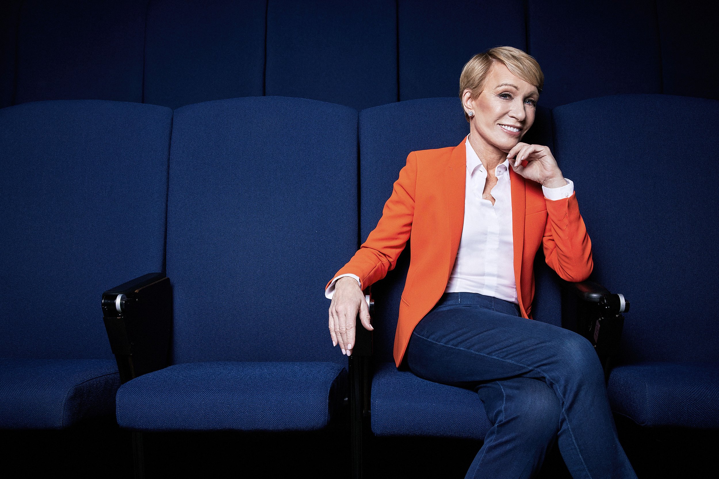 Portrait of Barbara Corcoran seated in an orange blazer against deep blue theater seating.