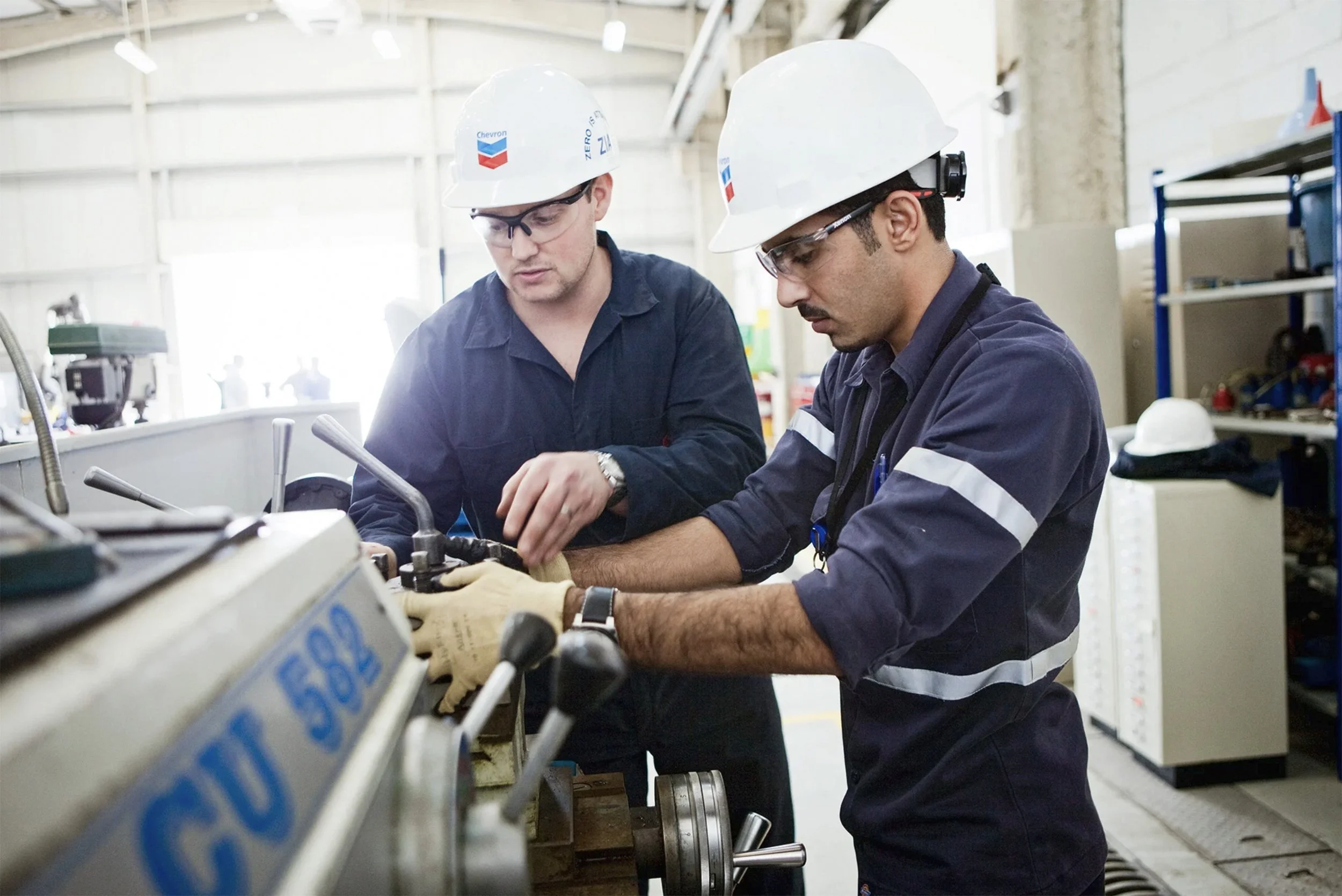 Two Chevron employees wearing hard hats work together on industrial machinery inside a facility.