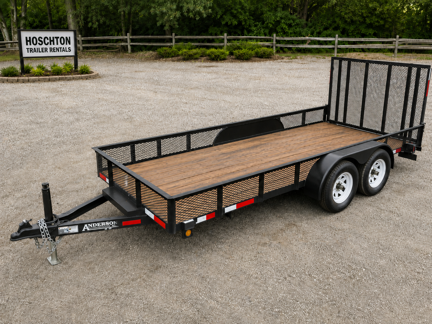 A flatbed utility trailer with a wooden deck and black metal frame parked on gravel. The trailer has two axles with white wheels and a hitch for towing, located near a sign that reads 'HOSCHTON TRAILER RENTALS'.