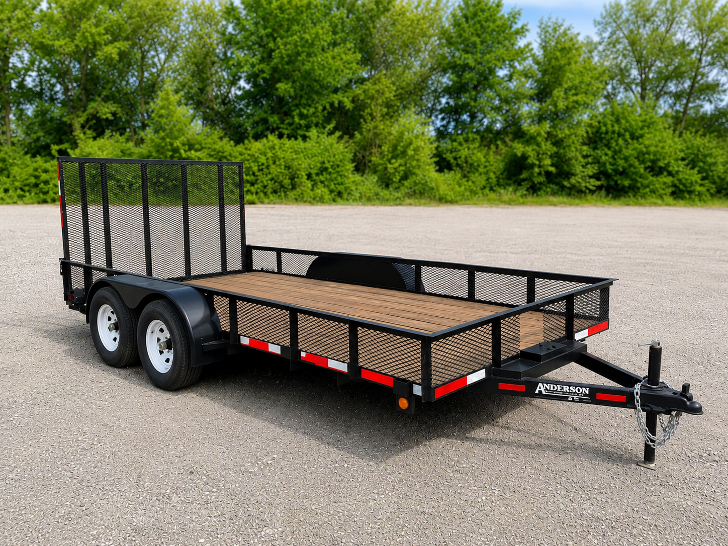 A black flatbed utility trailer with wooden floor and metal mesh sides parked on an asphalt surface with green trees in the background.
