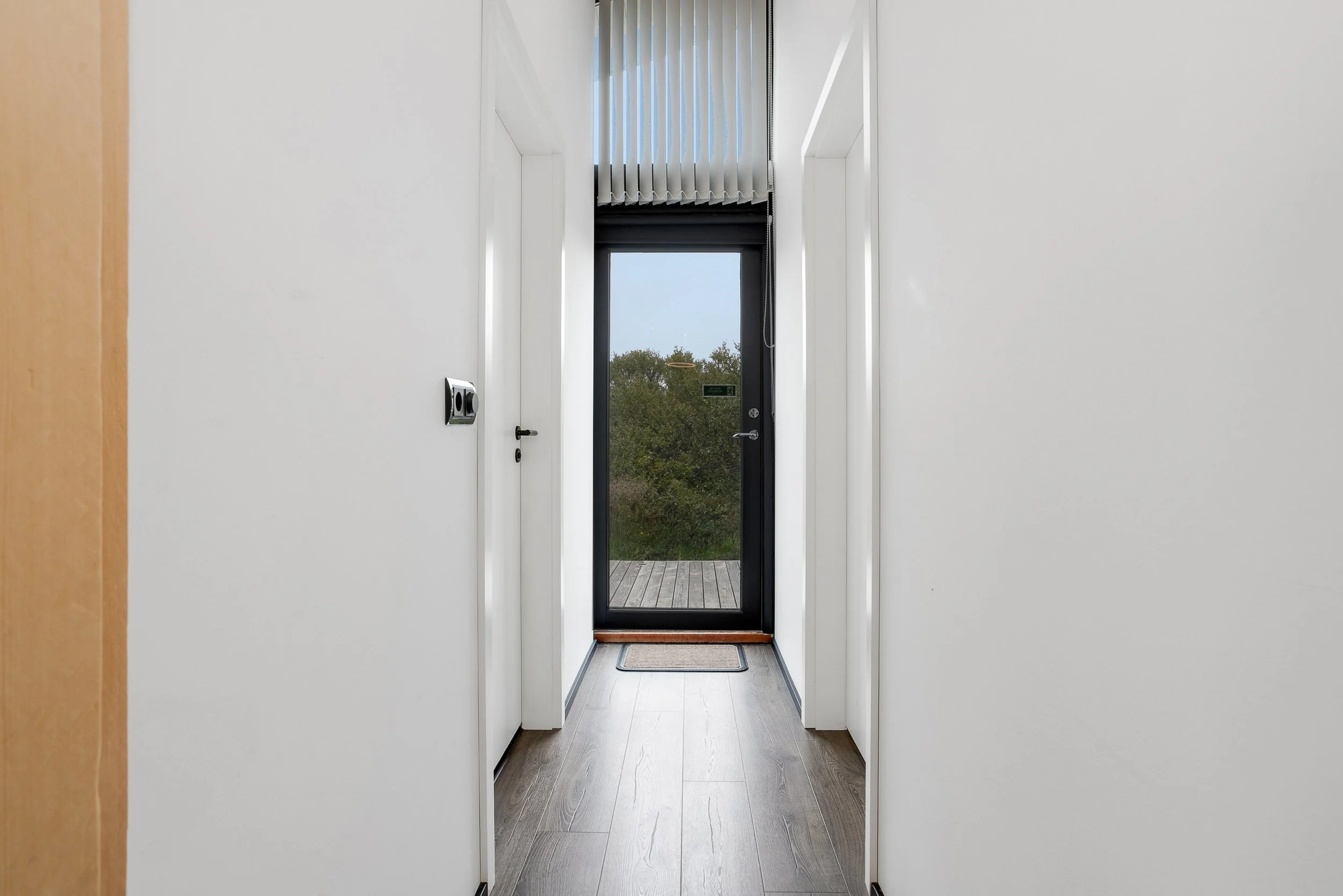 View of a hallway with white walls and a wooden floor, leading to a glass door with vertical blinds and a view of trees outside.