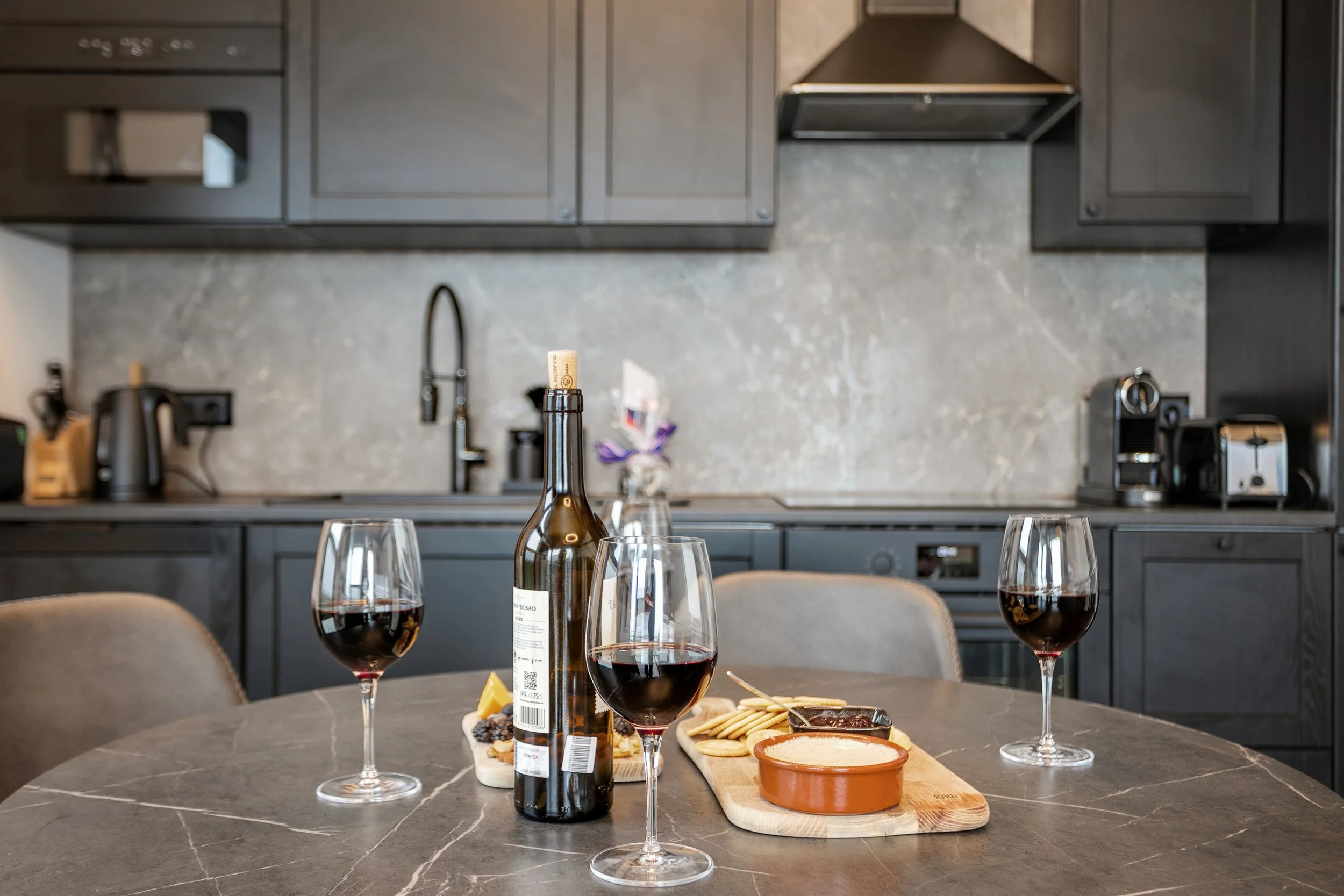 A kitchen countertop with three glasses of red wine, a wine bottle, and a cheese and cracker platter. In the background, there are gray cabinets, a gray backsplash, a sink, and kitchen appliances.
